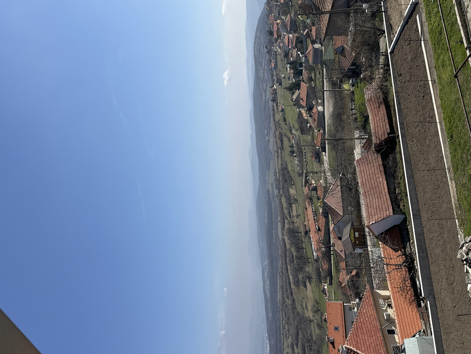 This image captures an elevated, wide view of the village of Chelopek, Bulgaria, under a clear, light blue sky with minimal, wispy clouds near the horizon. In the immediate foreground, a dark brown, tilled garden bed is visible, contained by concrete retaining walls. Green grass lines the area below and to the right of this bed. The midground features numerous houses with reddish-brown tiled roofs, typical of rural Bulgarian architecture, spread across undulating terrain. Bare deciduous trees, indicating late winter or early spring, are interspersed among the dwellings and in the surrounding fields, which show patches of green grass. Utility poles and wires are observable throughout the village. A paved road or dirt track winds through the residential areas. In the distant background, hazy rolling hills and mountains extend to the horizon. To the far left, beyond the main village cluster, faint structures suggest a more densely populated area or another town. No people are discernible, and no specific actions or interactions are visible within the frame.