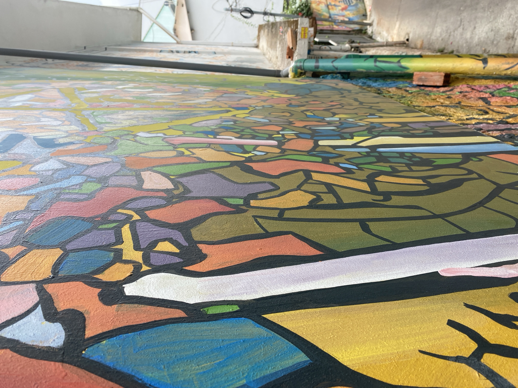 The image captures a section of an expansive, vividly colored mural in Da Lat, Vietnam, possibly a street art project. The mural features a patchwork of colors, shapes, and patterns delineated by thick black lines, reminiscent of stained glass. Shades of blue, green, yellow, orange, purple, and pink are prominent. The art is on a ground surface, indicating either a floor, pavement, or a very low wall.
In the background, elements suggesting the setting include a concrete structure, possibly a building facade or low wall, visible above the mural. Rolled-up segments of the artwork or painting materials are also present, supported by a brick. The overall environment suggests an outdoor or semi-outdoor urban space in Da Lat where artists are creating or displaying artwork. No people are visible in the frame.