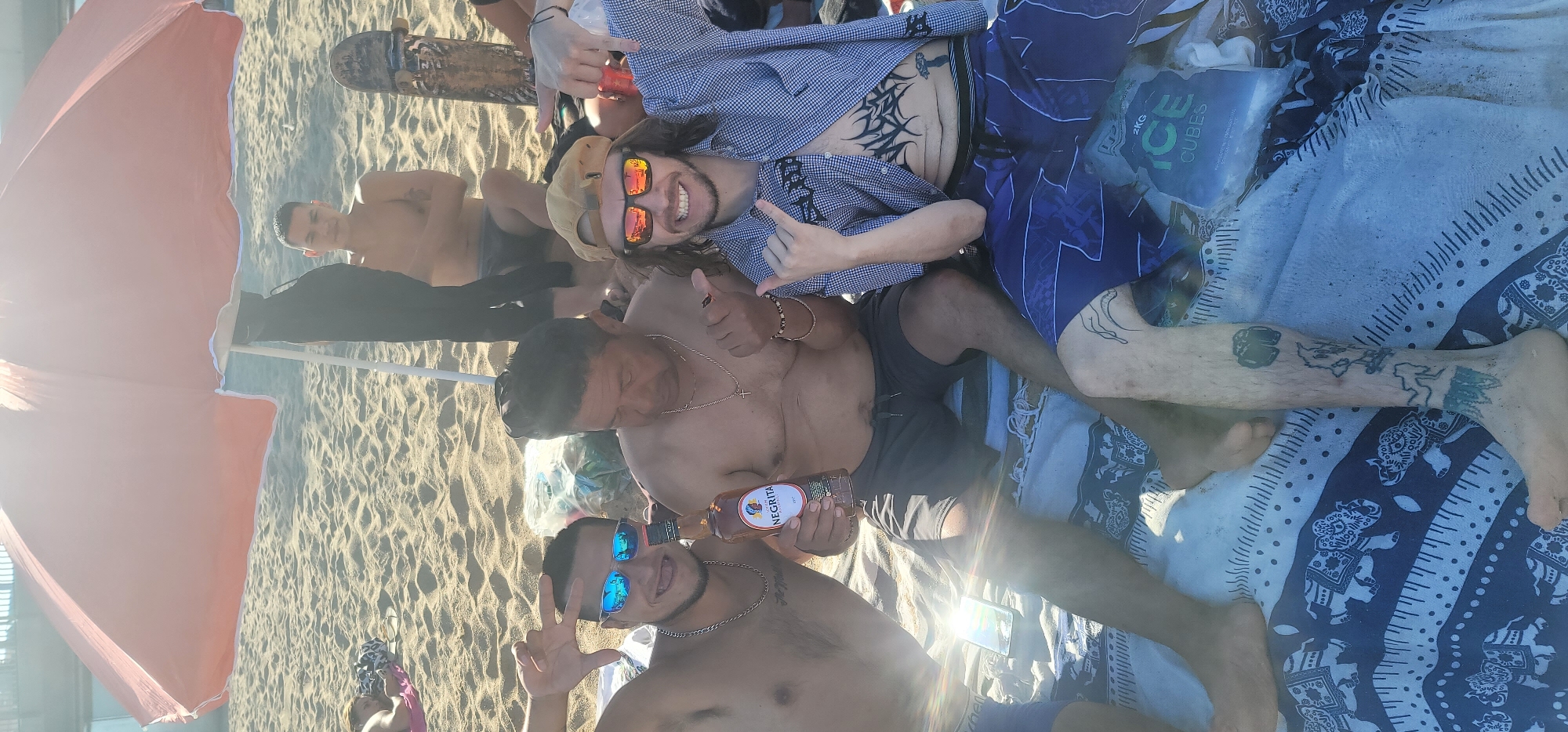 A full-length vertical photograph depicts a group of five individuals on a sandy beach under a large, peach-colored beach umbrella during bright daylight.
In the immediate foreground, three males are seated on a blue and white patterned blanket featuring elephant motifs. The male on the left, with dark hair and light brown skin, wears reflective blue sunglasses and a silver chain. He is shirtless, making a peace sign with his left hand and holding a dark glass bottle labeled "NEGRITA" rum in his right hand. The central male, an older individual with dark hair and medium brown skin, is shirtless, wearing dark swim shorts and a silver chain with a cross pendant; his gaze is directed downwards. The male on the right, with long brown hair, wears a beige cap reversed, reflective orange sunglasses, and an unbuttoned blue and white checkered shirt. A prominent black tribal-style tattoo is visible on his abdomen, and several other tattoos adorn his legs, including a wave-like design on his right thigh. He smiles broadly and makes "shaka" gestures with both hands towards the camera. A transparent plastic bag labeled "2KG ICE CUBES" rests on the blanket beside his right leg.
In the midground, partially obscured by the umbrella's pole and a black garment hanging from it, a fourth male with short dark hair and a small tattoo on his left bicep is seated, shirtless and wearing dark swim trunks. A skateboard with visible grip tape is leaning upright to his right. Further to the left in the midground, a fifth person is lying face down on a pink towel, largely obscured by the sand and sunlight.
The environment consists of light-colored sand under bright sunlight, which creates lens flare in the image. A faint, elevated structure, possibly a pier or walkway, is visible in the distant background above the horizon. The scene is a beach.