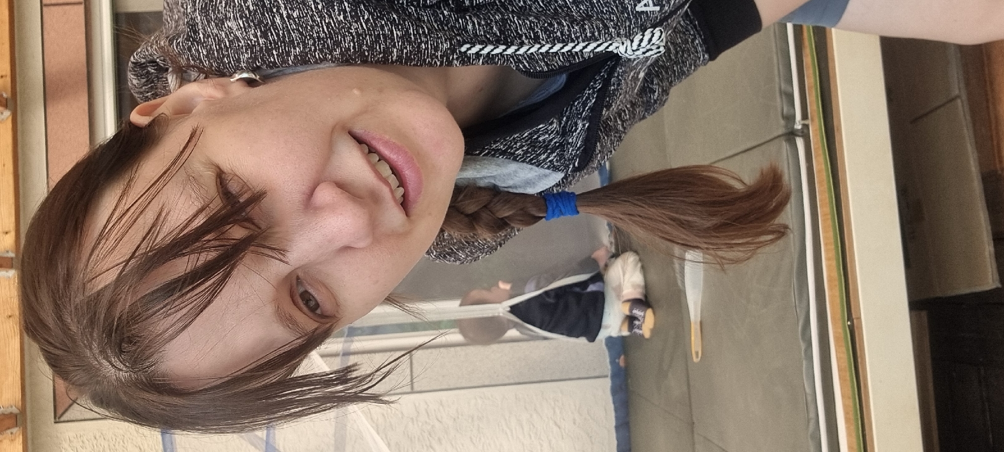 A woman with brown hair is photographed from a low angle, appearing to be smiling with her head tilted to her left. Her hair is styled with a braid secured with a blue hair tie. She is wearing a gray and white hoodie. The background includes what appears to be an open sliding glass door with a view of a building. Inside the home is a doll lying on the floor. The location in the city of Dunnae-myeon, South Korea is impossible to determine from the image alone.