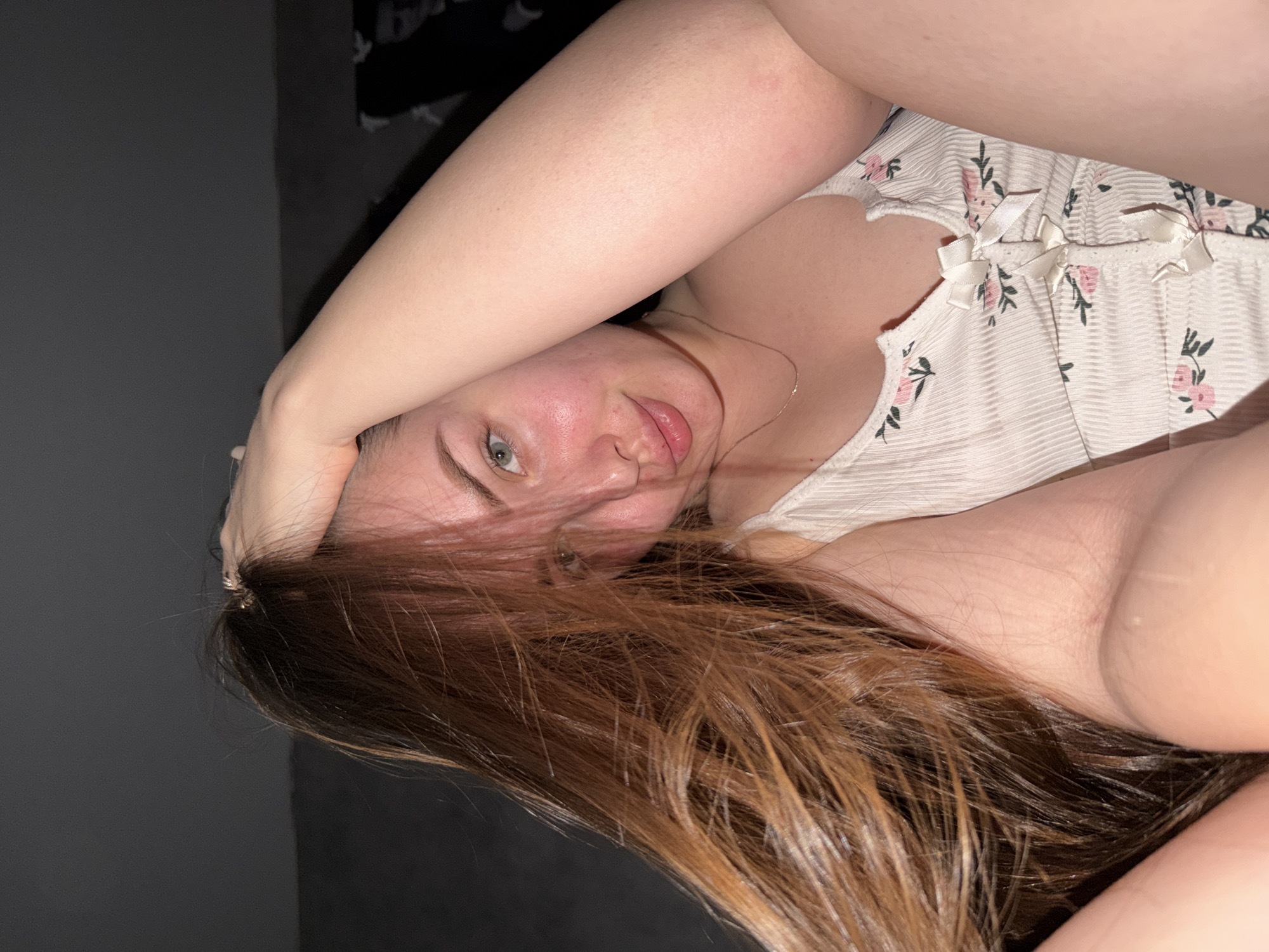 The image shows a woman lying down. She has fair skin, green eyes, and reddish-brown hair. She is wearing a light-colored top with a floral pattern and ribbon details. She has one arm raised above her head and is looking at the camera. The background is a dark gray wall. There are no other people or objects clearly visible. The location is unknown, but is speculatively the city of Chaikovsky, Russia.