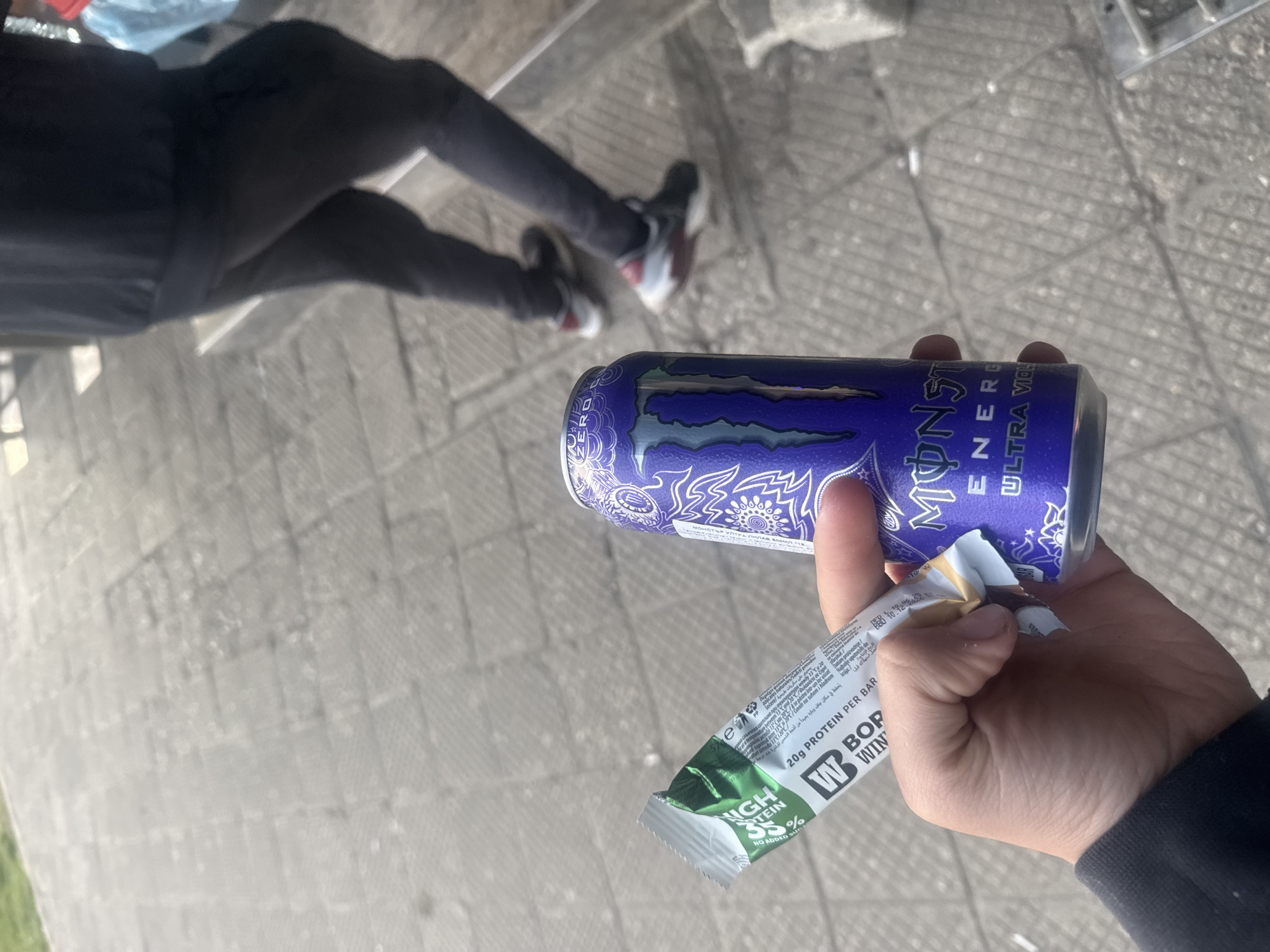 The image shows a person standing on a concrete surface, possibly a sidewalk or pedestrian area. Their legs, clad in dark pants and sneakers, are visible in the background. The person is holding a can of "Monster Energy Ultra Violet" and a "Born Winner" protein bar. The brand name is "Born Winner" and the flavor might be peanut caramel. The text on the packaging indicates it contains 20g of protein per bar.
There is no information in the image that explicitly indicates this is in Plovdiv, Bulgaria. The OCR text contains some Bulgarian, but that isn't enough to provide explicit certainty.