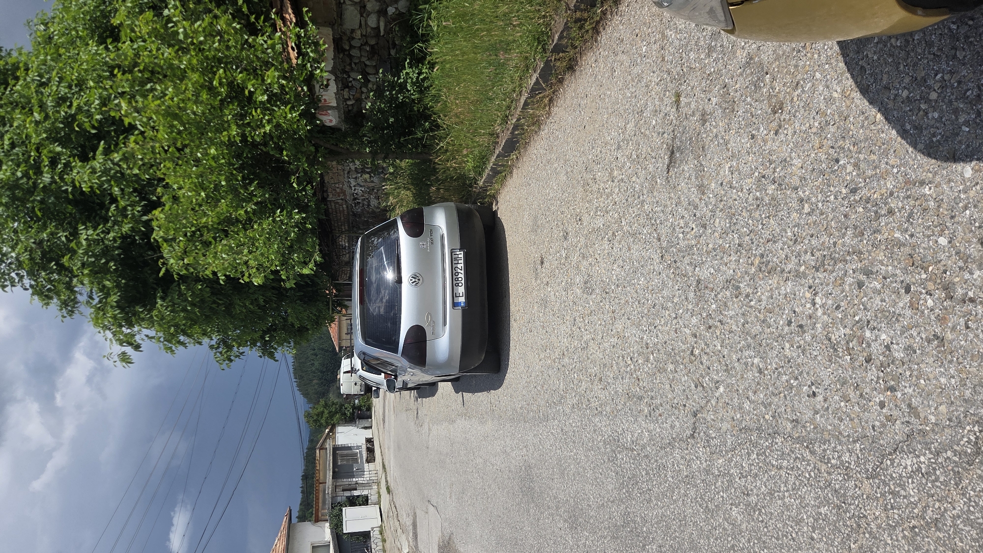 A silver Volkswagen Golf is parked on a paved road in a village. The car is facing away from the camera, and its license plate reads "E 8892 HH". A large green tree with dense foliage is to the right of the car, and a stone wall runs along the edge of the property behind the tree. In the background, there are houses and a forest-covered hill under a cloudy sky. A portion of a gold-colored car is visible in the lower right corner of the image.