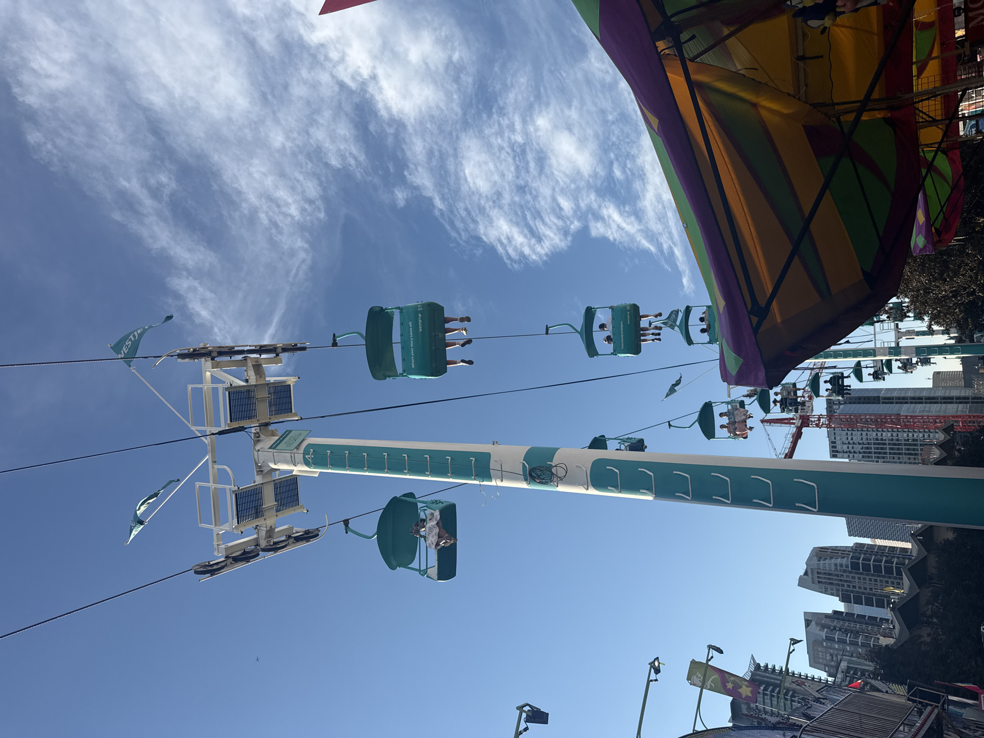 A low-angle, wide shot captures a Ferris wheel with teal gondolas against a bright blue sky with wispy clouds. Several gondolas are visible, each carrying passengers. The Ferris wheel structure itself is tall and white with teal stripes. In the background, a city skyline with numerous modern buildings, including high-rises, is visible, suggesting an urban environment. A red construction crane is also present amongst the buildings. A portion of a colorful tent structure, with purple, green, and yellow panels, is in the foreground on the right. The image was taken in the city of Calgary, Canada, likely at a fair or amusement park during daylight.