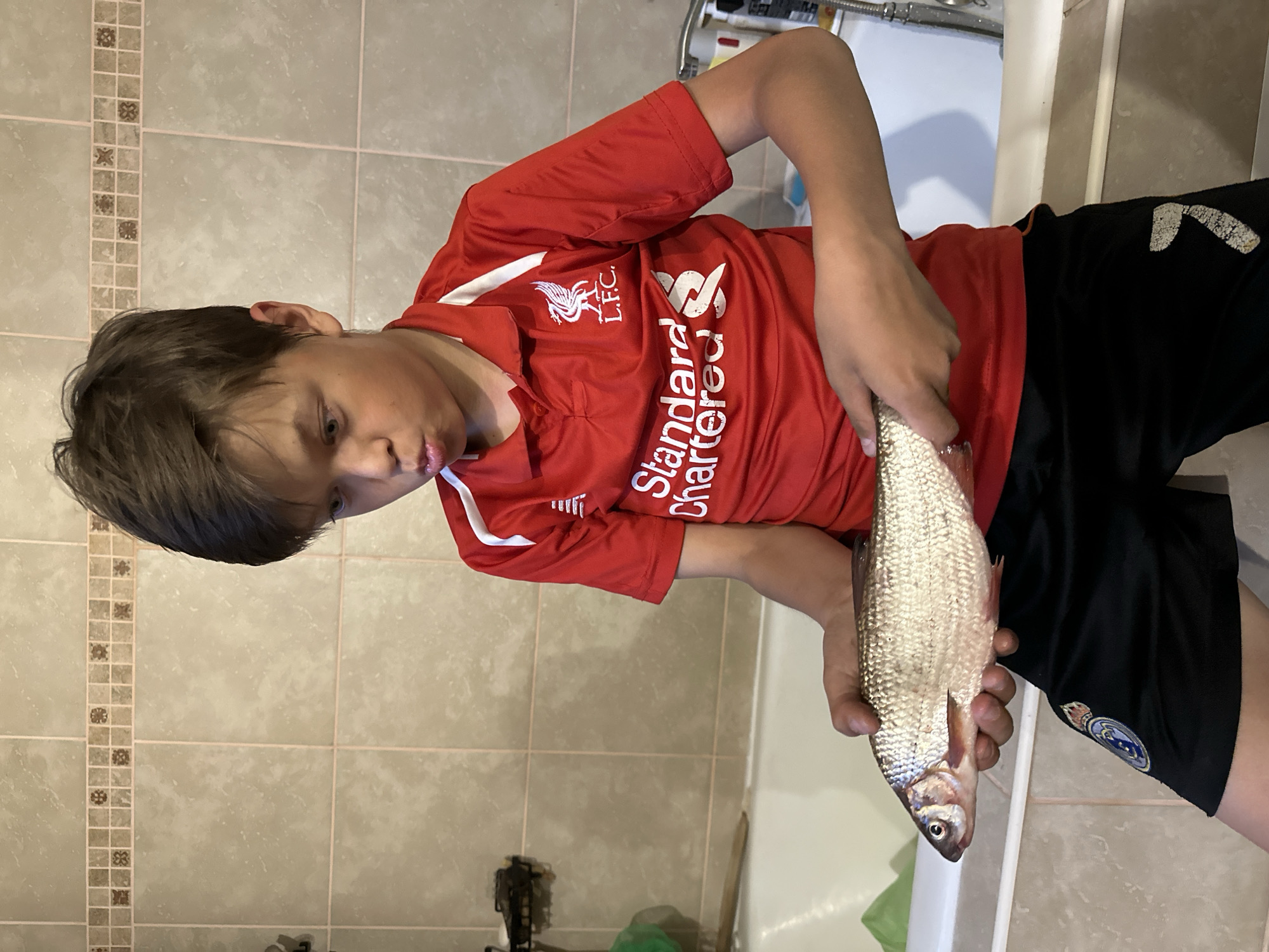 A young male is shown holding a fish indoors. The boy is wearing a red Liverpool soccer jersey and black shorts with a Real Madrid logo visible. The background consists of tiled bathroom walls and a bathtub. The location is inferred from the original request, which is Chaikovsky, Russia.