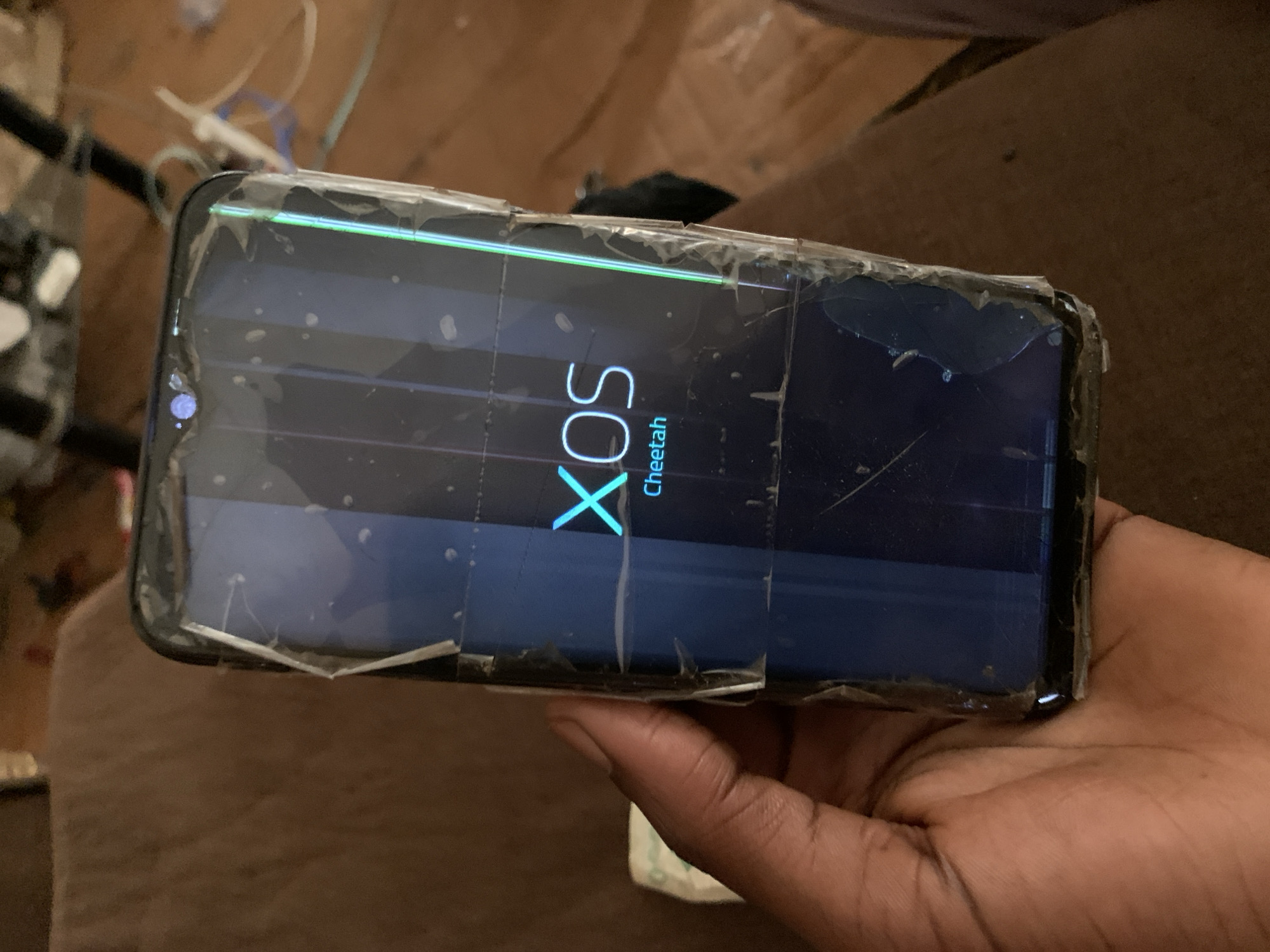 A person's hand is holding a cracked smartphone. The phone's screen displays the Infinix logo, with the text "XOS Cheetah" beneath it. The screen also shows vertical lines of light, indicating damage. The background is blurry and appears to be a brown, textured surface, possibly a couch. There are also some out-of-focus objects in the background, including what looks like wires and paper. The image was taken in Jos, Nigeria.