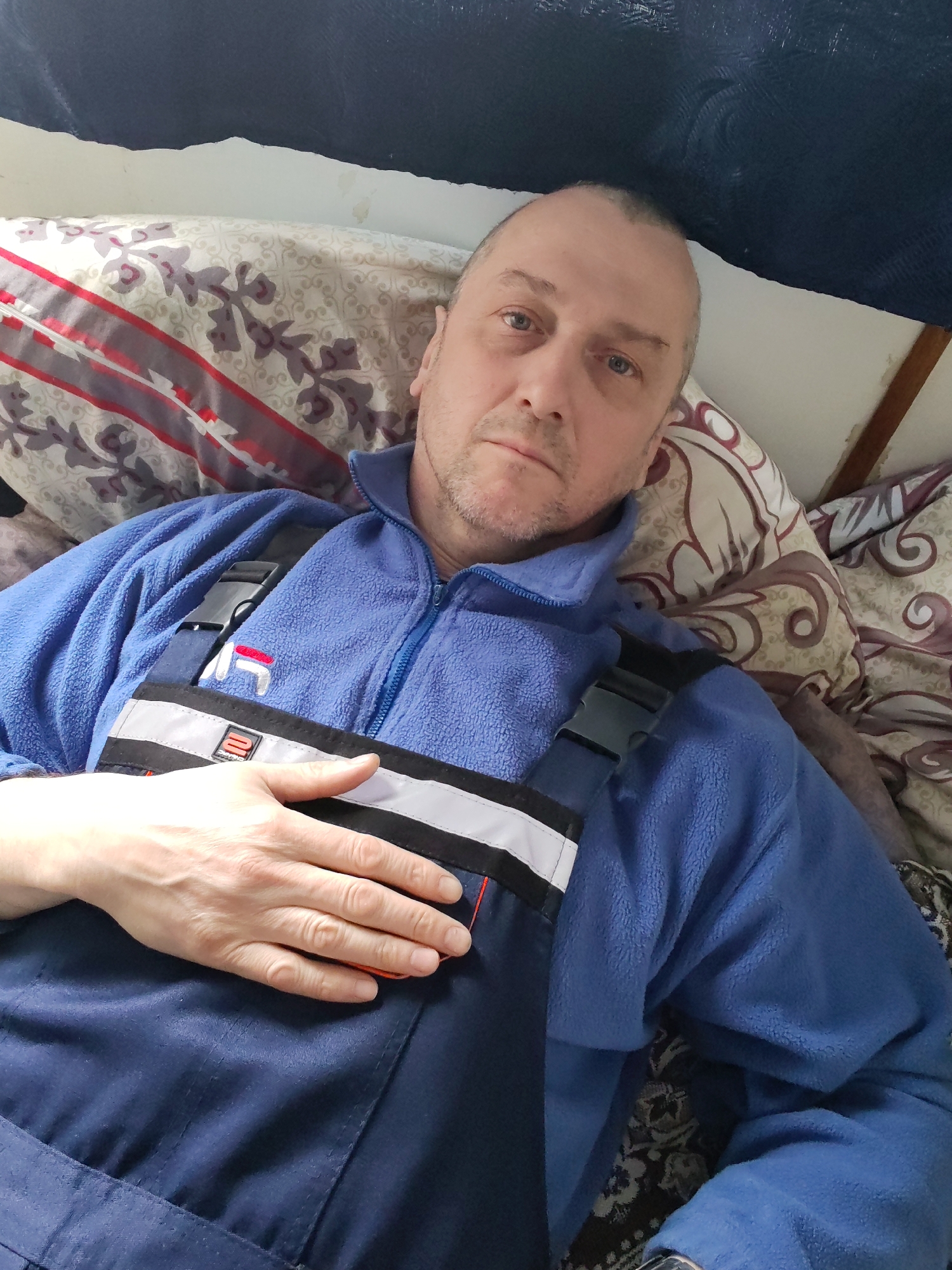 A man lies in an unknown indoor location, appearing to be in a bed or on cushions, with his head near a bed frame or wall. He is wearing a blue fleece jacket over a blue work overall with reflective stripes across the chest. His right hand rests on the overall. The background consists of patterned pillows and a dark-colored surface, possibly a headboard. His facial expression is neutral or slightly tired.