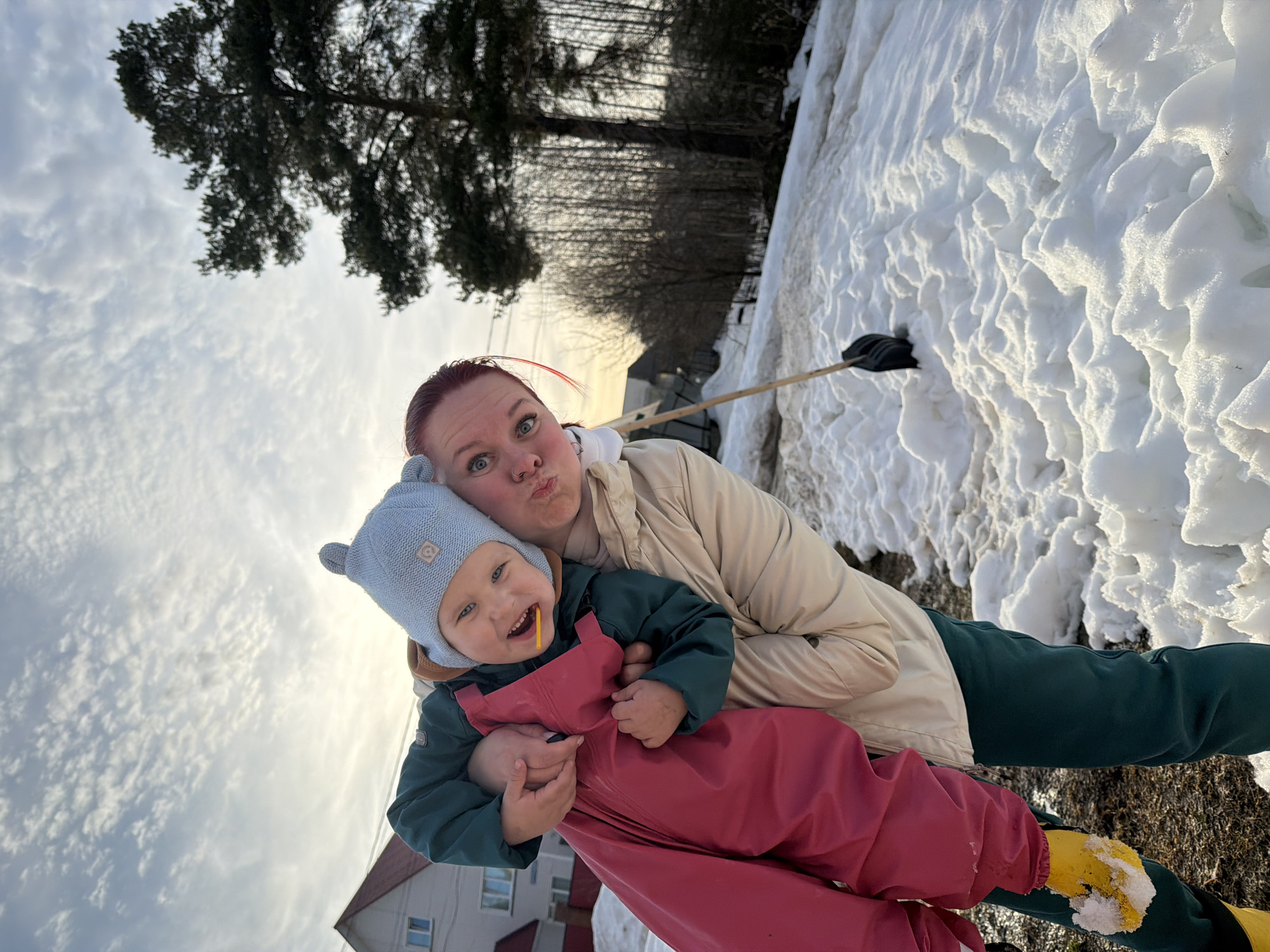 An adult female and a young child are captured in an outdoor setting in Chaikovsky, Russia, characterized by melting snow and clear skies with scattered cirrus clouds. The adult, with red hair and a visible nose piercing, wears a light beige puffer jacket and dark green pants, and is holding the child securely. She gazes towards the viewer with a puckered lip expression. The child, dressed in a light blue knitted hat with ear-like protrusions, a dark green jacket, and red overalls over yellow boots, smiles broadly, revealing a small yellow object held between the teeth. The ground is covered unevenly with white snow and exposed patches of dirt. In the mid-ground, to the right, a black shovel with a wooden handle is embedded vertically in a snowdrift. The background features a two-story residential building with a reddish roof and white facade on the left, and a dense stand of trees, including a prominent evergreen conifer on the right, and numerous leafless deciduous trees further back. The overall lighting suggests either morning or late afternoon.