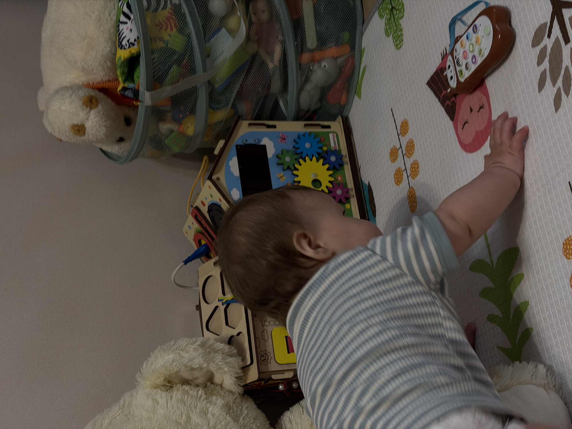 A baby in a blue and white striped shirt is reaching towards an electronic toy on a patterned playmat. The baby is positioned near a busy board and a stuffed animal. A toy storage container is also present. The location appears to be indoors, possibly a child's room.