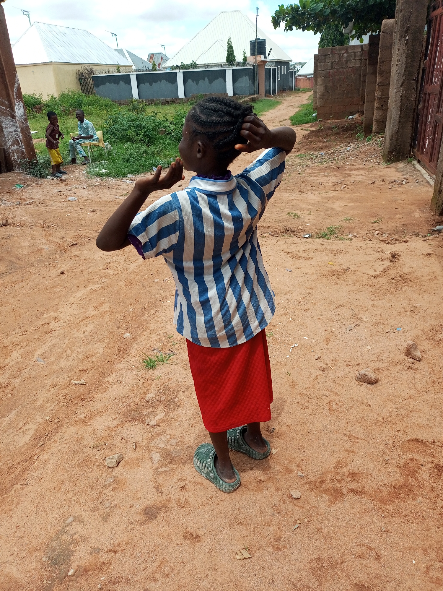 In Tempure, Nigeria, a young Black girl stands with her back to the camera in a dirt pathway, her hands touching her braided hair. She wears a blue and white striped short-sleeved shirt and a red skirt. Her feet are clad in green rubber clogs. Two other figures, a child and an adult, are visible in the background, seated amongst lush green vegetation near a row of houses with white roofs. The environment appears to be a residential area with brick walls and a weathered wooden gate on the right side of the frame. The ground is sandy with scattered stones and dried leaves, indicating an outdoor setting.