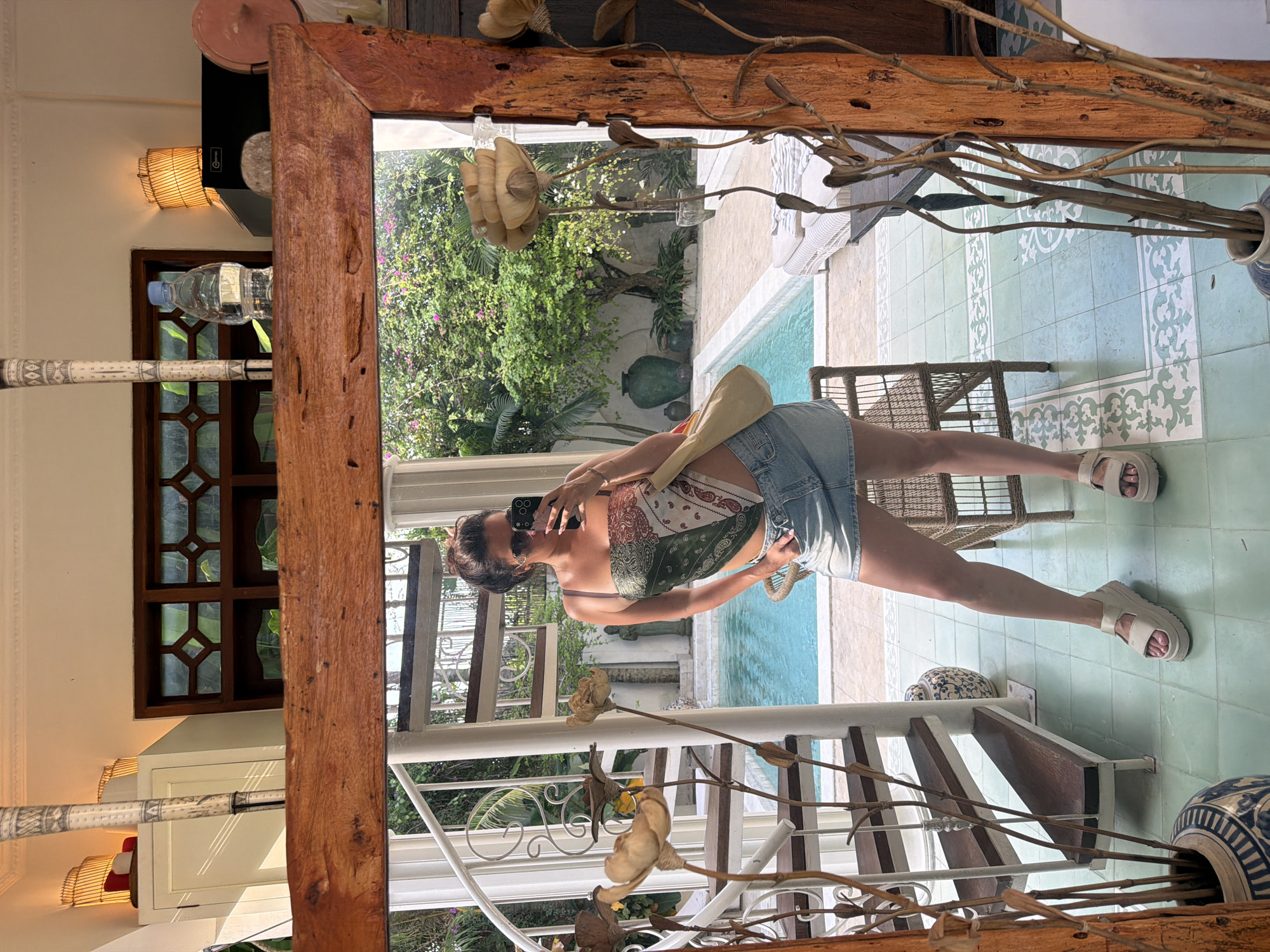 A woman takes a mirror selfie in a room with a wooden-framed mirror, reflecting an outdoor swimming pool and lush greenery. She is wearing a bandana-style top, a denim skirt, and platform sandals. The room features a spiral staircase, decorative vases, and natural light filtering in from the outdoor space. The setting appears to be a tropical villa in North Kuta, Indonesia.