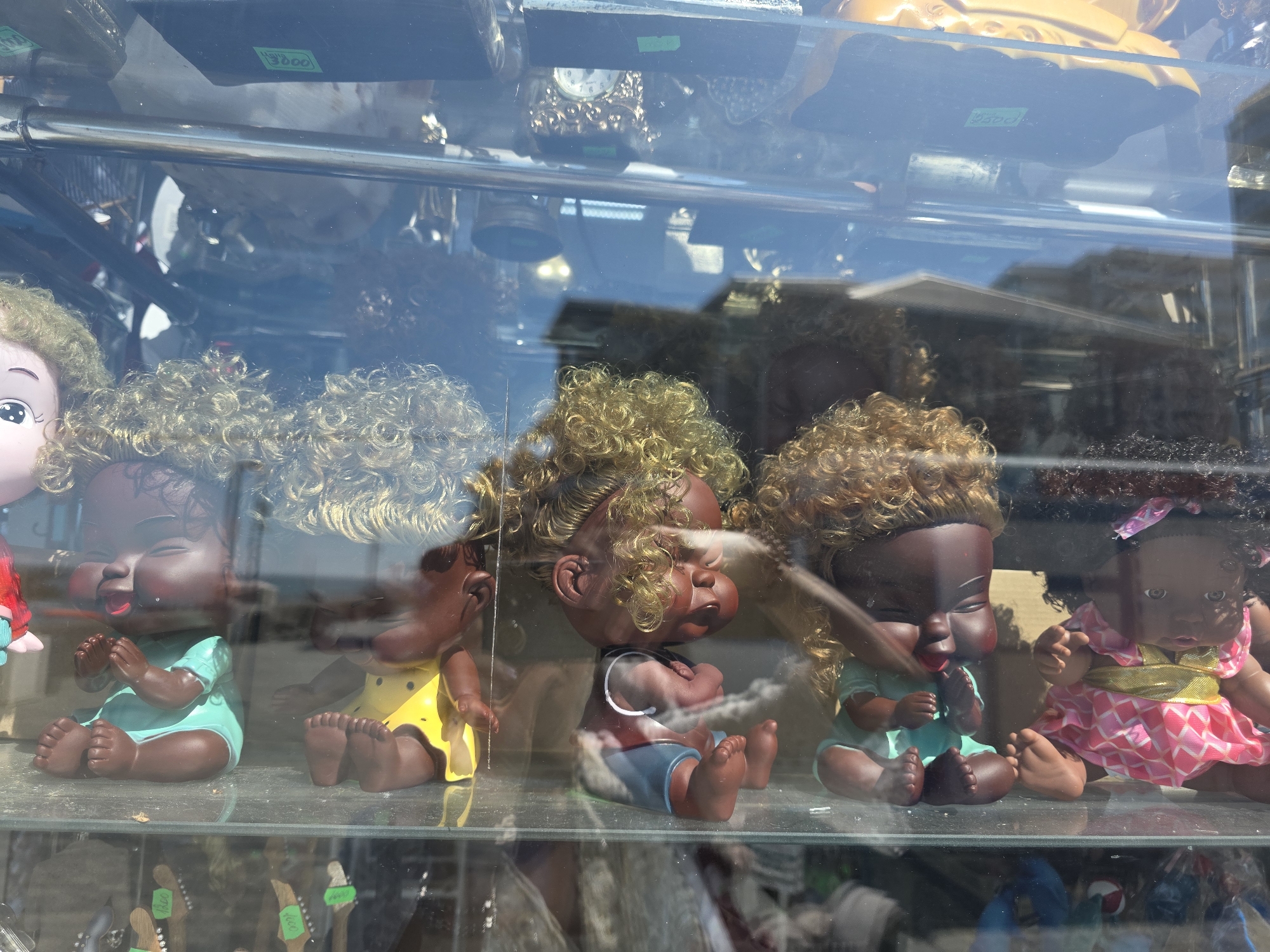 The image displays a retail store window in Sochi, Russia, under bright daylight, featuring a row of five dark-skinned dolls on a clear glass shelf. From left to right, a partially visible doll with blonde curly hair is followed by a full doll in a light blue outfit, seated with clasped hands and a joyful, closed-eyed expression with puffed cheeks. Next, a doll in a yellow outfit exhibits a similar cheerful, puffed-cheek expression. A third doll wears a red top and blue shorts, with arms crossed and a pouting, closed-eyed, puffed-cheek expression. The fourth doll, dressed in light blue, mirrors the first full doll's pose and expression. The final doll on the right has dark curly hair with a pink bow, wearing a pink patterned dress, with open eyes and hands slightly raised.
A horizontal chrome bar runs across the upper section of the display. The glass reflects a clear blue sky, a distant horizon that appears to be the sea, and sections of nearby buildings. Above and through the display glass, various retail items are visible on shelves, including a decorative clock, a gold-colored ornate object, and several green price tags bearing numbers such as "1800", "3800", and "1000," indicative of Russian Ruble prices. Reflections and visual penetration through the lower part of the glass reveal more merchandise on a shelf below the dolls, including items resembling miniature guitar necks with additional price tags ("1000", "1800"). The scene suggests a souvenir or general merchandise shop located in a sunny, outdoor area of the coastal city.
