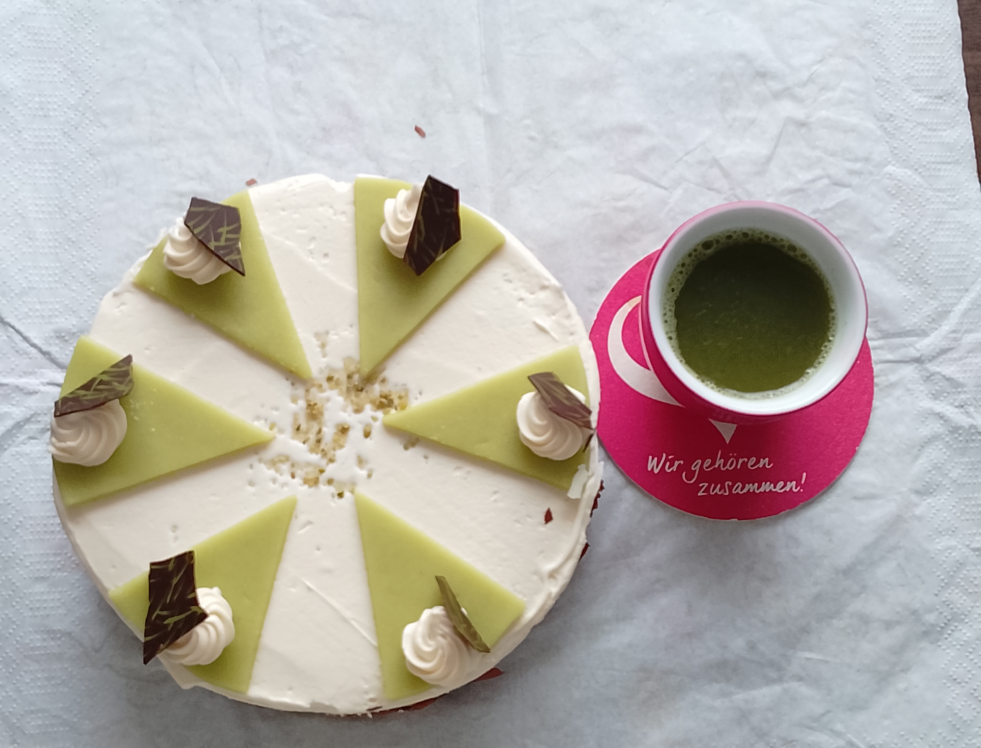 A white frosted cake with sliced green triangular toppings and dollops of white cream with chocolate shavings is displayed on a white textured surface. Adjacent to the cake is a pink cup filled with a green beverage, possibly matcha tea, resting on a pink circular coaster with white text. The coaster reads "Wir gehören zusammen!" which translates to "We belong together!". The setting appears to be indoors, on a table or counter, as suggested by the crumpled white paper napkin or tablecloth. There are no individuals visible in the image.