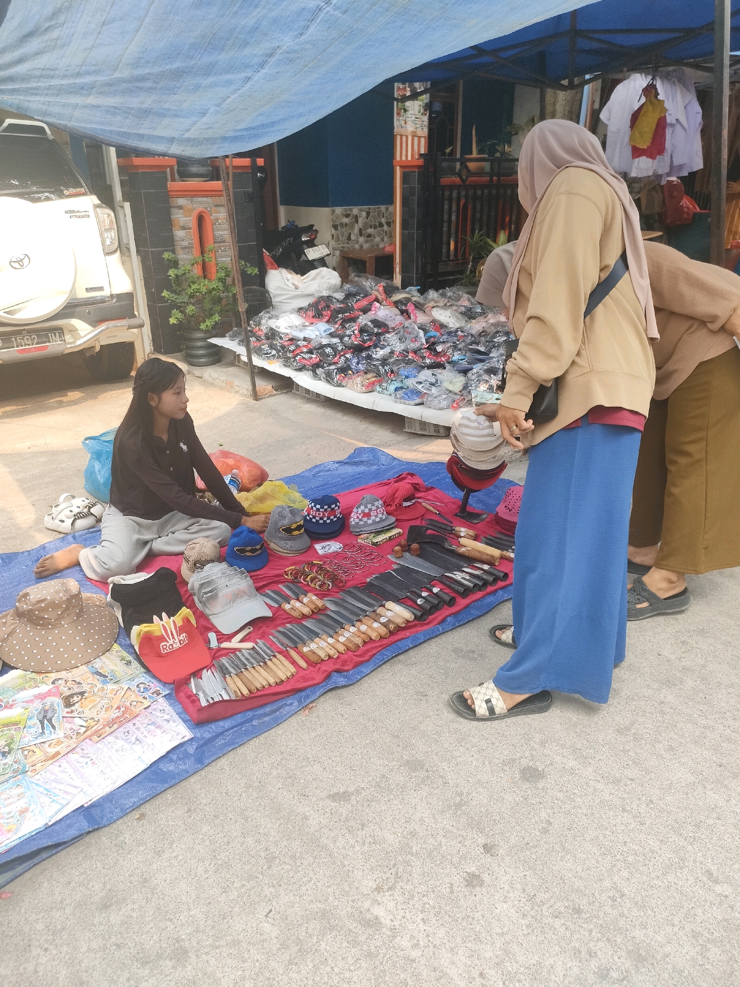 The image depicts a street scene in Batam, Indonesia, likely a market stall. A young woman is seated on a blue tarp spread on the ground, surrounded by various items for sale. To her left are a white car with a spare tire mounted on the back, and a potted plant. In front of her, also on the tarp, are several hats, including a wide-brimmed polka-dot hat, a red cap with "Rabbit" written on it, and various knitted and baseball caps. Adjacent to the hats is a collection of knives displayed in a row, with wooden handles. Behind the woman and the merchandise, a stall structure is visible, draped with a blue tarp overhead. Piles of folded clothing and other goods are stacked on shelves or tables. Two other individuals, partially obscured, are standing near the stall, appearing to interact with the merchandise. One person, wearing a beige hoodie and a light pink hijab, is looking at the items on display, while another person in a brown sweater is facing away from the camera, also near the stall. The ground is paved with concrete. The overall atmosphere suggests a busy outdoor marketplace.