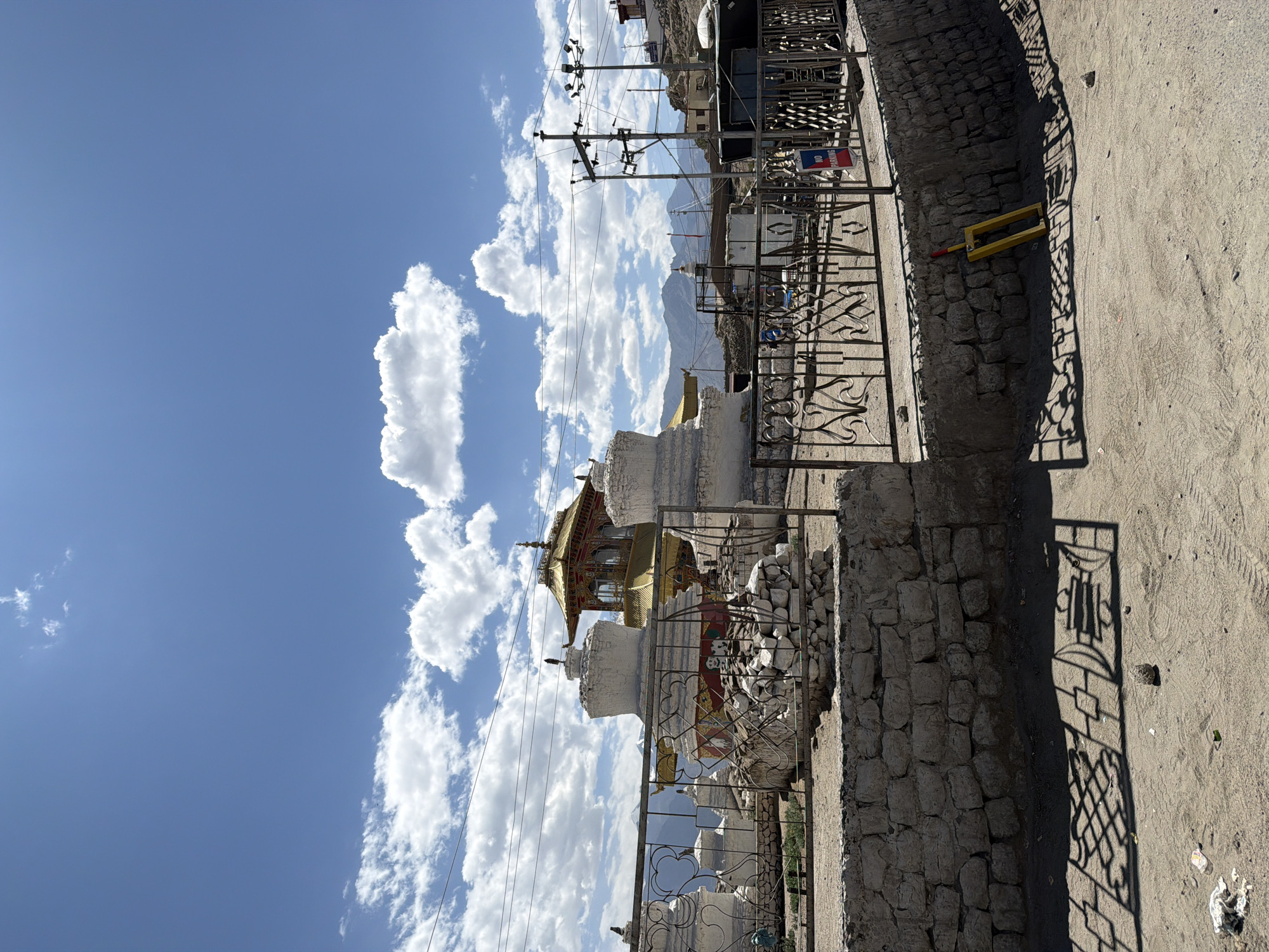 In the city of Leh, India, an outdoor scene features a religious complex with several stupas and a golden-roofed structure. A metal fence encloses part of the complex. The foreground shows a dirt ground with tire tracks and shadows cast by the fence. A yellow object, possibly a parking stopper, rests against a stone wall. In the background, mountains are visible under a blue sky with scattered clouds. Power lines and poles are present on the right side of the image. No people are visible in the frame.