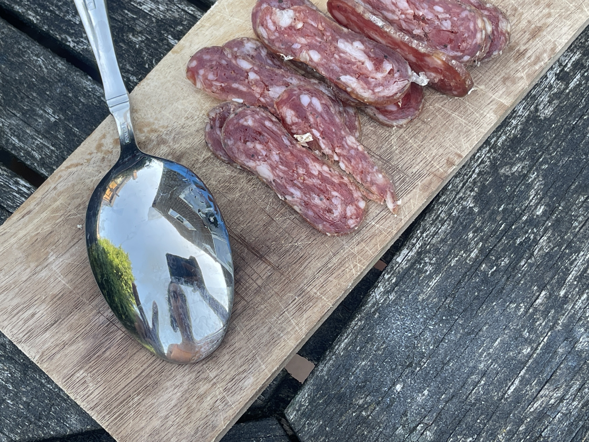 On a wooden surface, likely an outdoor table, a wooden board displays several slices of salami alongside a large silver spoon. The spoon reflects the surroundings, including a building and green foliage. The setting appears to be outdoors, possibly in a garden or patio in Maidstone, United Kingdom, based on the architectural style visible in the spoon's reflection. The overall scene is a casual food arrangement.