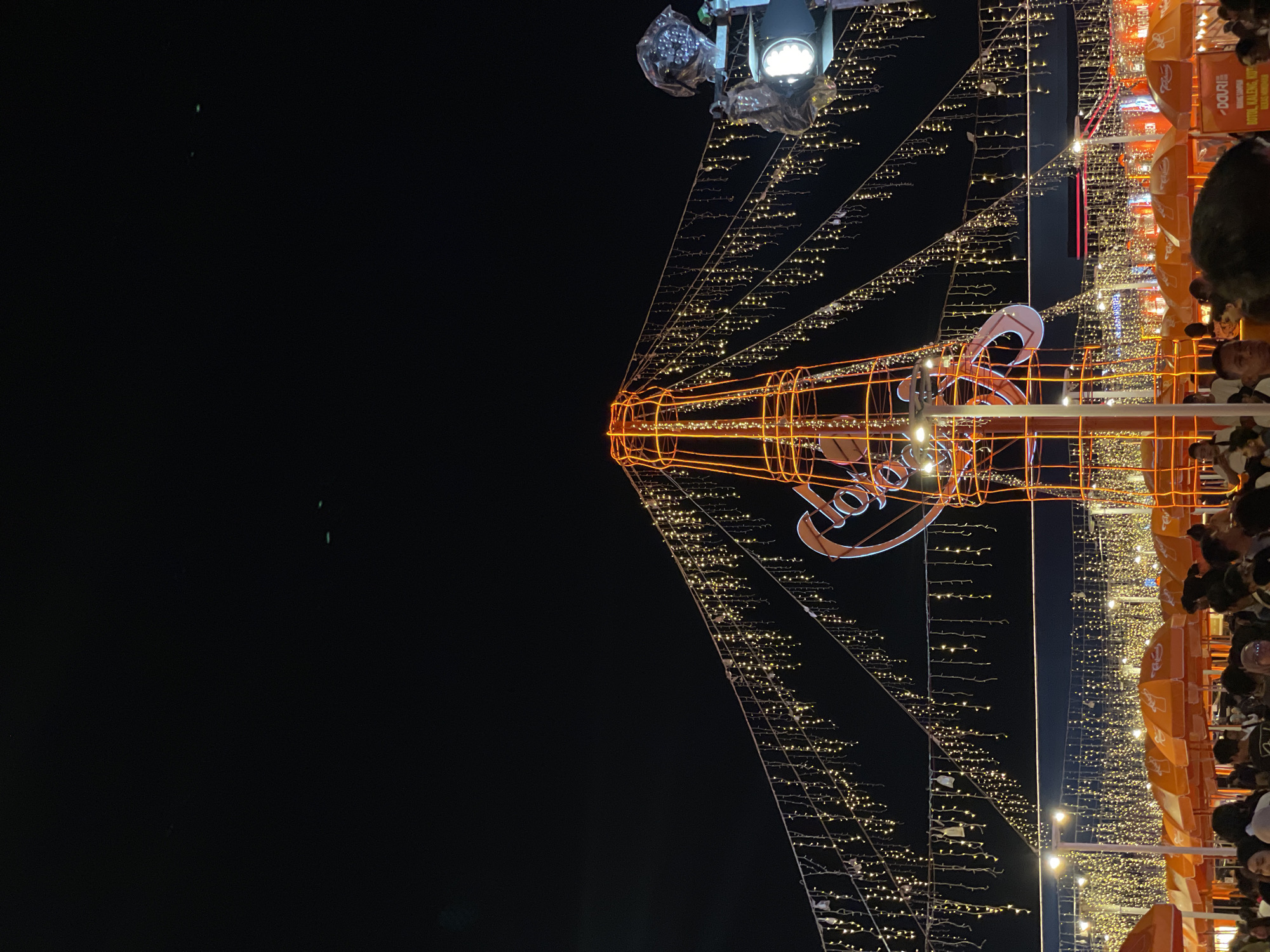 The image, taken at night in Salembaran, Indonesia, depicts an elaborate light display. A large, stylized bottle-shaped structure, outlined in orange lights, dominates the scene. The word "COLOR," also rendered in orange lights, is integrated into the design. Above the bottle-shaped structure, a lighting rig is visible. Strings of lights radiate outwards and downwards from the central structure, creating a canopy effect. To the right side of the image, a dense crowd of people can be seen, partially obscured, near what appear to be orange-colored stalls or awnings, also illuminated with strings of lights. The backdrop is a dark night sky.