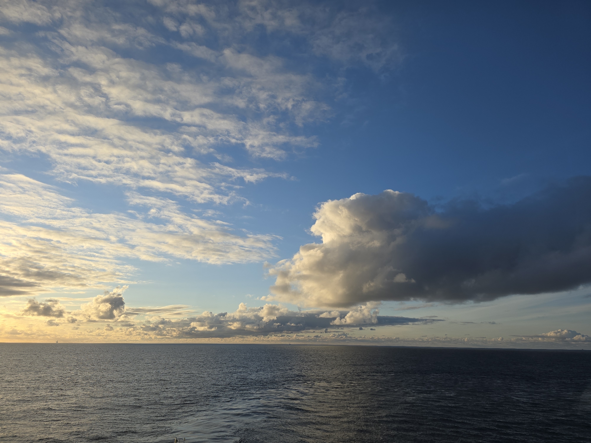 The image shows an ocean scene under a cloudy sky. The ocean is a dark blue color, with gentle ripples on the surface. The sky is a mix of light blue and dark blue, with scattered clouds. Some of the clouds are illuminated by the sun, while others are dark and ominous. The horizon line is visible in the distance, with a hint of land. There are no people or other objects visible in the scene. The image appears to have been taken from a high vantage point, such as a ship or cliff.