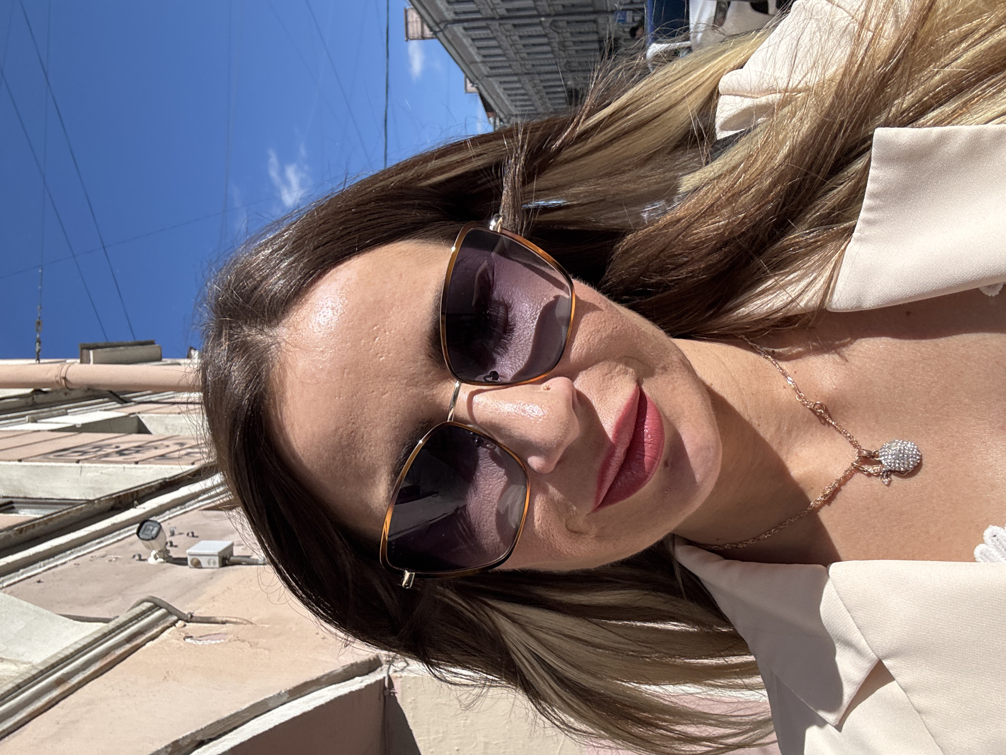 This is a close-up, selfie-style shot of a woman in Saint Petersburg, Russia. The woman is wearing sunglasses and has on a light-colored blazer. She has long, wavy brown and blonde hair. A delicate necklace with a sparkling pendant is visible around her neck.
The scene is outdoors on a sunny day, suggested by the bright light and shadows. The background includes a clear blue sky with some wispy clouds, overhead wires, and the facade of a building, likely in an urban setting. The angle of the shot, looking upwards, hints at a location with buildings. The woman's expression appears relaxed and she seems to be enjoying the daylight. The overall mood is casual and bright.