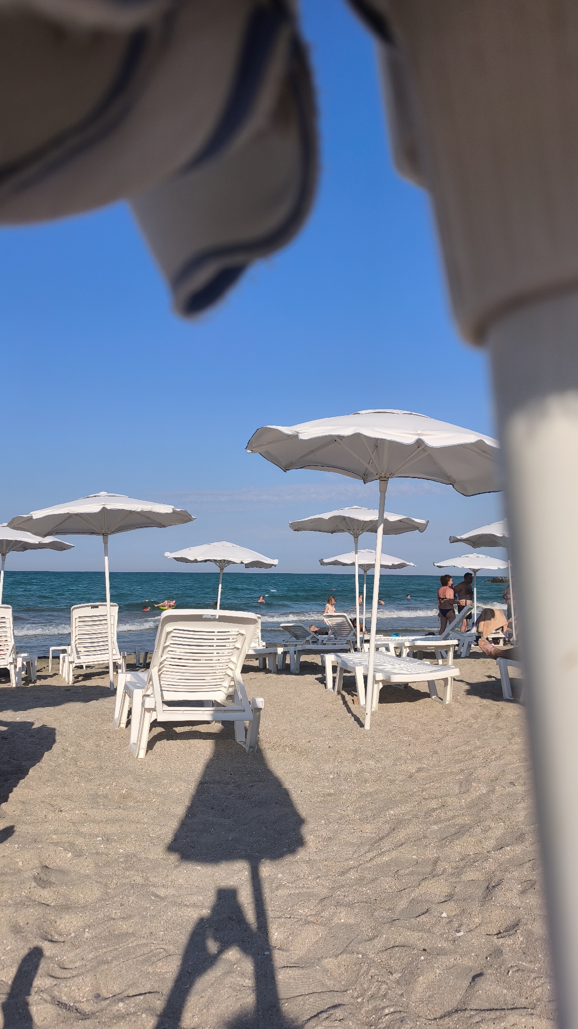The image captures a sunny day on a sandy beach in Pomorie, Bulgaria, with the sea visible in the background. The foreground is dominated by the sand and the shadow of a beach umbrella and a person holding a phone, suggesting the photo was taken by a tourist.
Scattered across the sand are numerous white plastic beach chairs and white beach umbrellas, most of which are open and appear ready for use. Some of these chairs are occupied by people, while others are empty.
In the middle ground, the calm, blue sea stretches to the horizon under a clear blue sky. Several individuals are visible in the water, some swimming or floating on inflatable devices. Along the shoreline, more beach chairs and umbrellas are arranged.
Further back, a few more beach chairs and umbrellas are visible. There are groups of people standing and sitting on the sand, some appearing to be engaged in conversation or relaxing. The overall impression is a typical scene at a popular beach resort. The presence of the beach chairs, umbrellas, and people enjoying the water indicates it is a pleasant day for a beach outing.