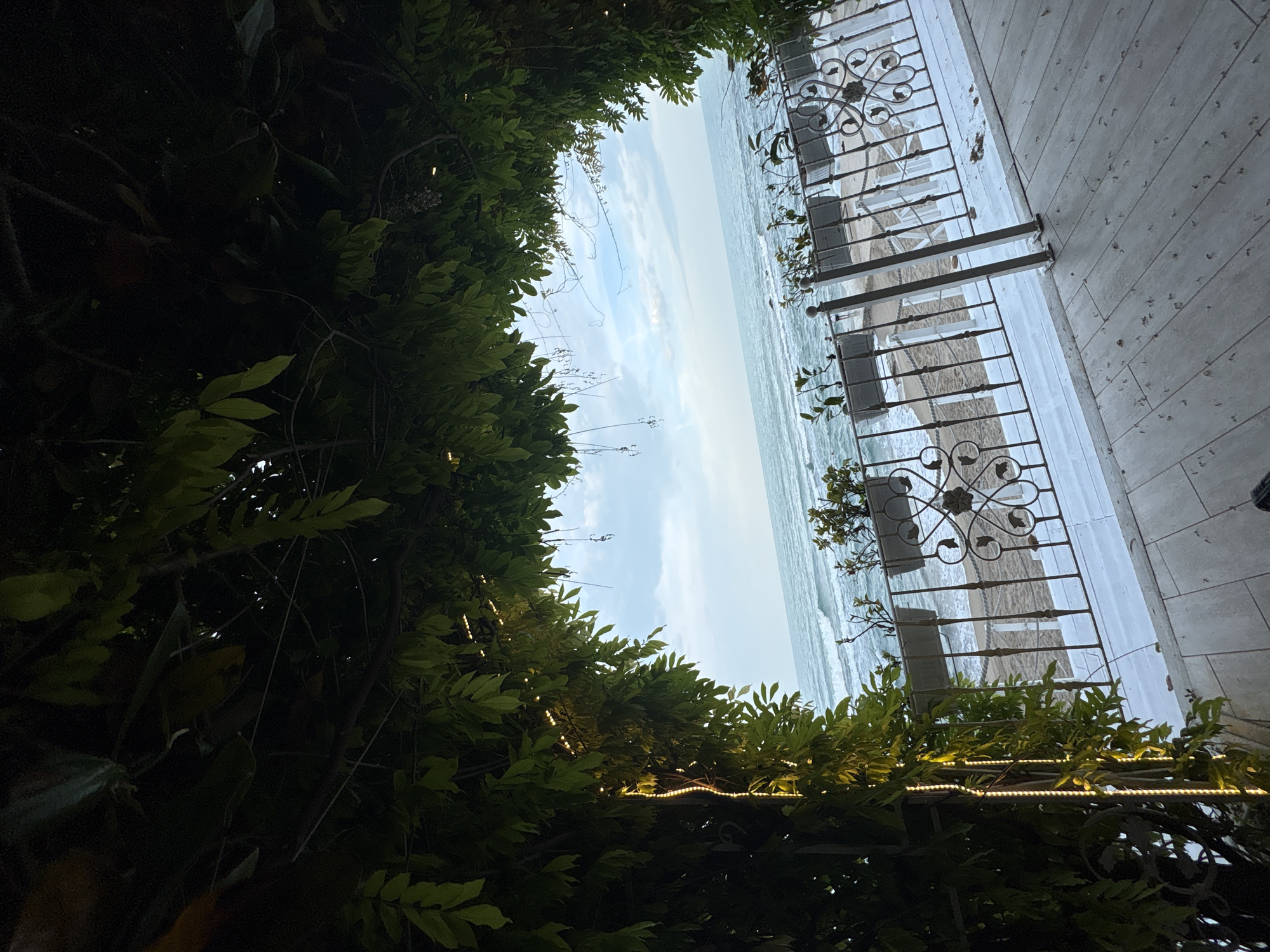The image displays an outdoor scene, likely a balcony or terrace, overlooking the sea. The view is framed by lush, dark green foliage along the top and bottom. Along the bottom foliage, small warm-toned string lights are visible, casting a soft glow.
Through the natural frame, a wide expanse of ocean is visible, with noticeable waves and whitecaps under an overcast, light blue sky. To the right, a tiled floor or patio with light-colored, rectangular tiles extends towards a decorative metal railing. This railing features intricate, ornate scrollwork. Beyond the railing, a sandy beach area can be seen leading to the water, and several light-colored chairs and tables are visible, suggesting an outdoor dining or seating area.
No people are present in the image. The string lights, combined with the overcast sky, suggest either late afternoon/early evening ambiance or a cloudy daytime setting. The scene evokes a tranquil, seaside atmosphere. There is no visible text.