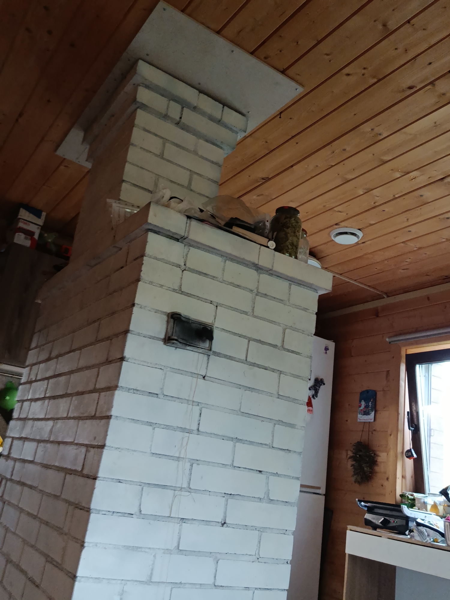 The image shows an interior view of a room, likely a kitchen or living area, in a wooden house. A prominent white brick structure, resembling a fireplace or a stovepipe, dominates the left side of the frame. This structure extends upwards towards a wooden ceiling. The ceiling is made of horizontal wooden planks, with visible beams. On the right side of the image, a white refrigerator is visible, and further to the right, a window looks out onto an unseen exterior. A countertop with various items, including jars and a small appliance, is also present. The overall impression is of a rustic, domestic setting. The location is identified as Verea, Russia.