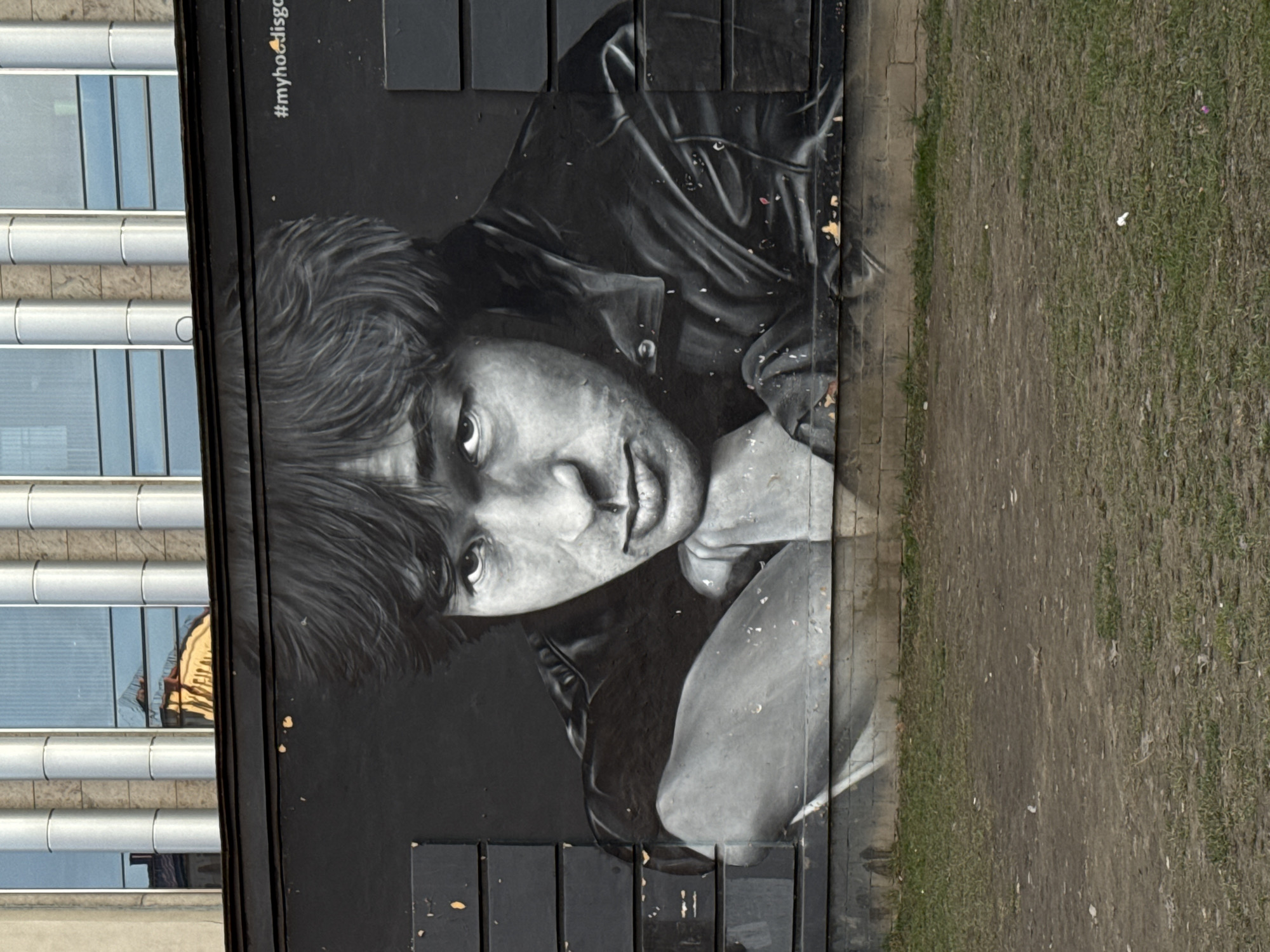 The media file displays an outdoor scene, dominated by a large, monochrome mural painted on a dark vertical wall. The mural features a detailed, realistic portrait of a person, likely male, with shaggy dark hair and striking eyes, looking directly at the viewer. The subject is depicted wearing a dark, possibly leather, jacket, with one hand resting near their chin or mouth area, conveying a contemplative or intense expression. The artwork is executed in grayscale, utilizing shades of black, white, and grey to create depth and contrast.
To the left of the mural (when viewed in its natural upright orientation), a modern building is visible, characterized by multiple floors with reflective glass windows and prominent horizontal metallic bands. These windows show reflections, including a faint image of what appears to be a person in a yellow jacket. To the right of the mural, the ground consists of a narrow dirt path running alongside a patch of somewhat sparse, dry-looking grass. The wall itself shows some minor wear and tear, with small chips and marks visible in places.
In the upper right section of the mural, a hashtag is visible: "#myhoodisgo". The overall lighting suggests it is daytime. The image, although provided in a rotated format, depicts a vibrant piece of street art integrated into an urban environment, located in Saint Petersburg, Russia.
