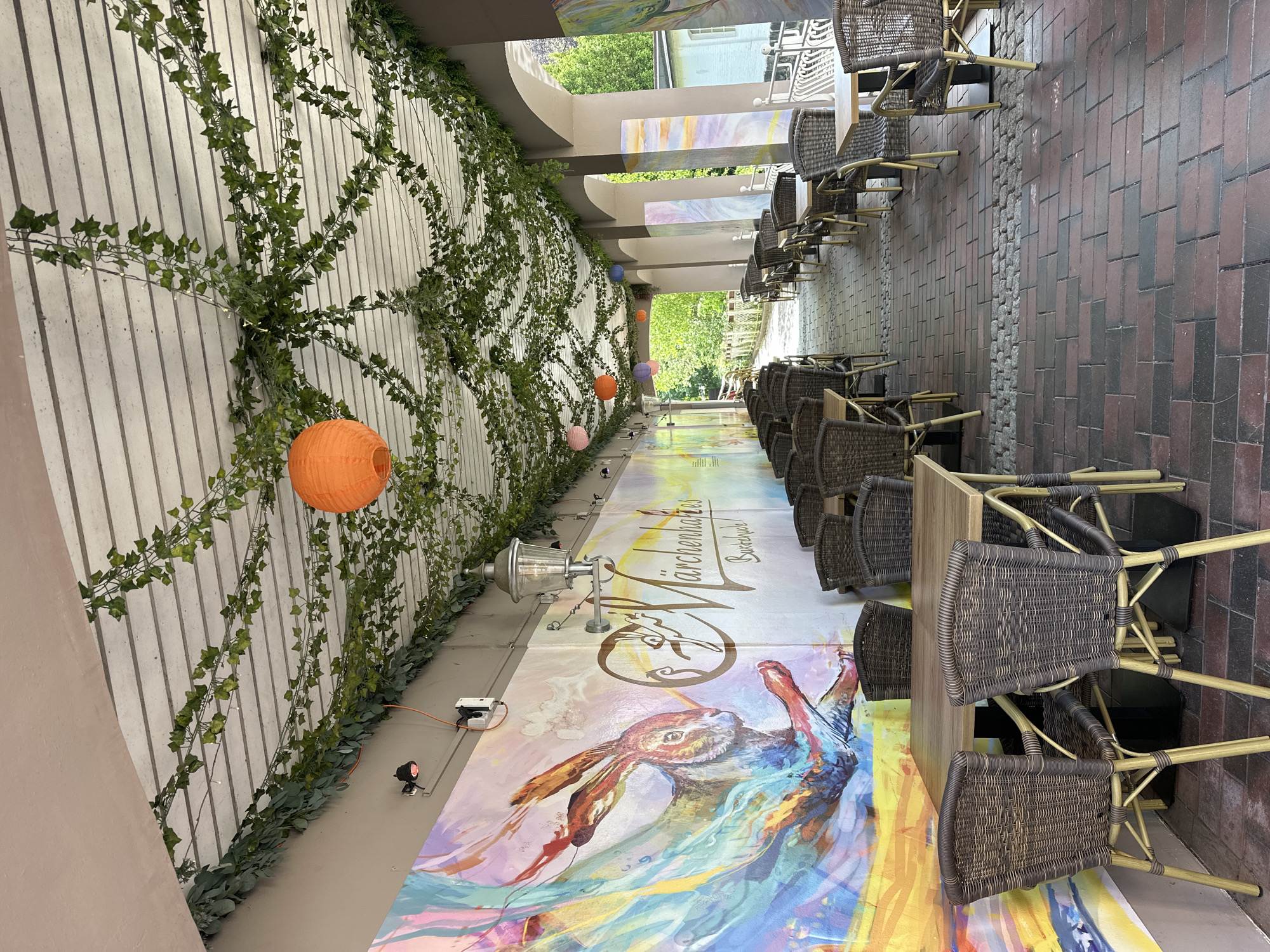 The image shows an outdoor seating area of a café or restaurant, located in Buxtehude, Germany. The seating area has tables and wicker chairs. The ground is covered with a painted mural of a hare. A white wall runs along one side, decorated with ivy and orange paper lanterns. Lights are placed on the ground. On the opposite side, the paving is made of bricks. Beyond the seating area, there is a view of greenery.