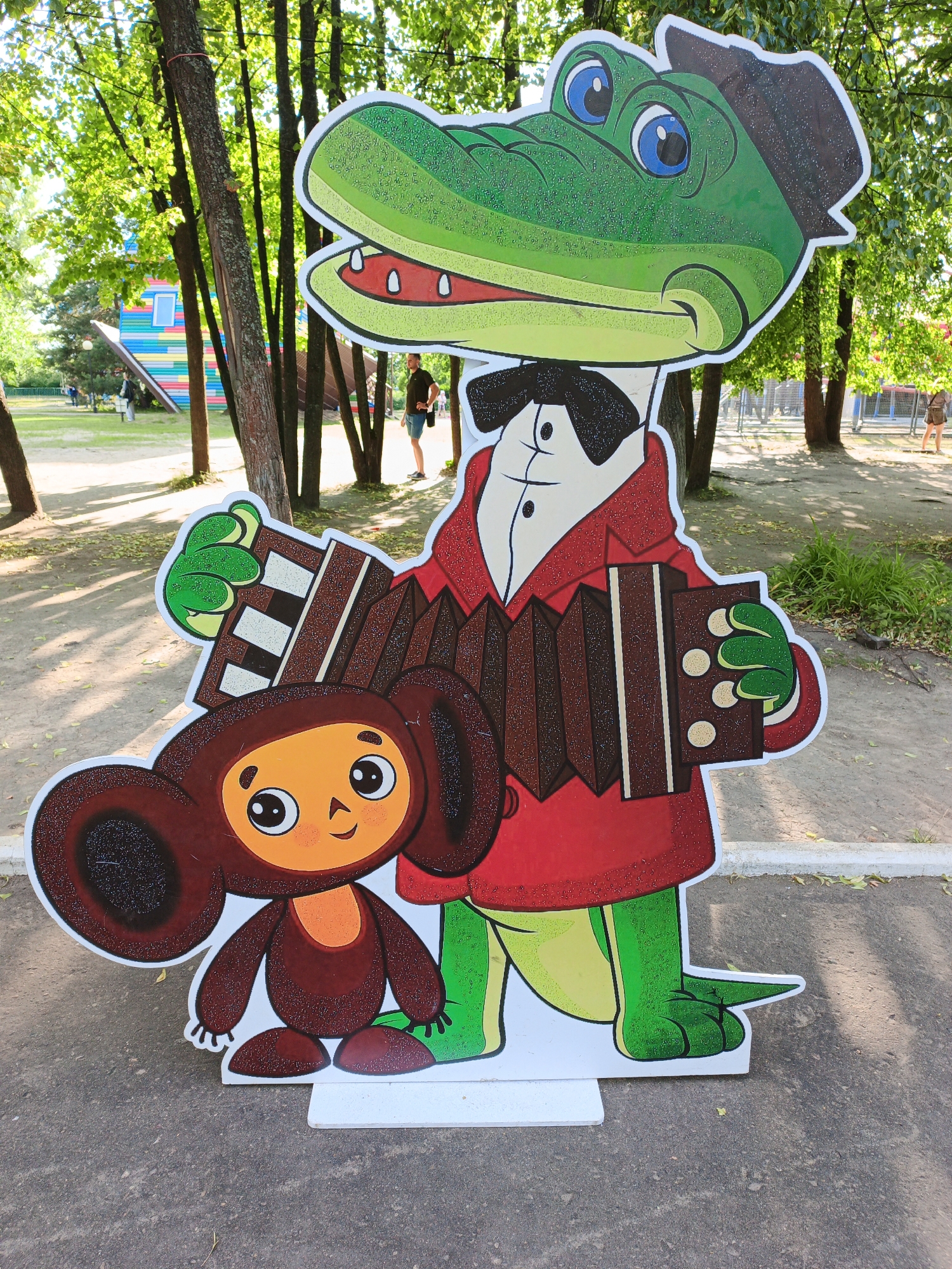 A cardboard cutout of two cartoon characters, a crocodile and a small brown creature, stands outdoors on a paved surface. The crocodile, dressed in a red coat and a hat, is holding an accordion. The small creature, with large ears and a round body, is positioned in front of the crocodile. Both characters are brightly colored and appear to be smiling.
In the background, trees with green leaves suggest a park or outdoor recreational area. Sunlight filters through the trees, creating dappled shadows on the ground. In the distance, there are hints of structures, including a colorful, tilted building and some fencing. A few people can be seen walking in the background, indicating the area is open to the public. The overall atmosphere seems relaxed and cheerful, suggesting a sunny day. There is no visible text in the image.