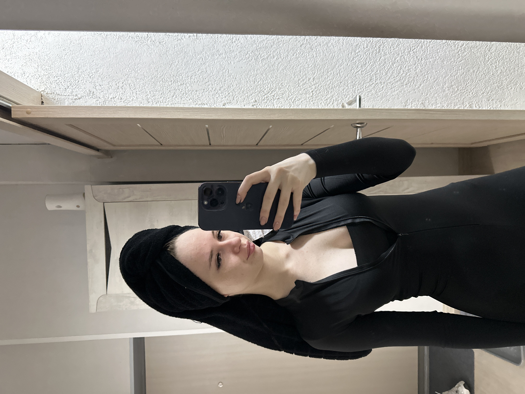 A woman is taking a selfie in an indoor bathroom setting. She is wearing a black long-sleeved outfit with a zipper at the chest and a black towel wrapped around her head. She is holding a dark-colored smartphone in her right hand, using it to capture the image. The background includes a bathroom vanity with a light-colored countertop, a textured white wall, and a cabinet above the vanity. The location is unknown.