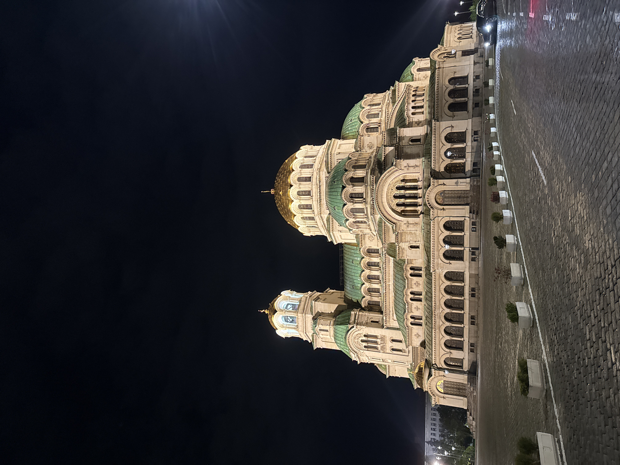 The image shows the Alexander Nevsky Cathedral in Sofia, Bulgaria, at night. The cathedral's architecture is prominent, with its gold domes and green roofs illuminated against the dark sky. A cobblestone street runs alongside the cathedral, marked with white lines and decorative planters. A car is parked on the street near the building. The sky is dark and clear, with light sources causing some flares near the top.