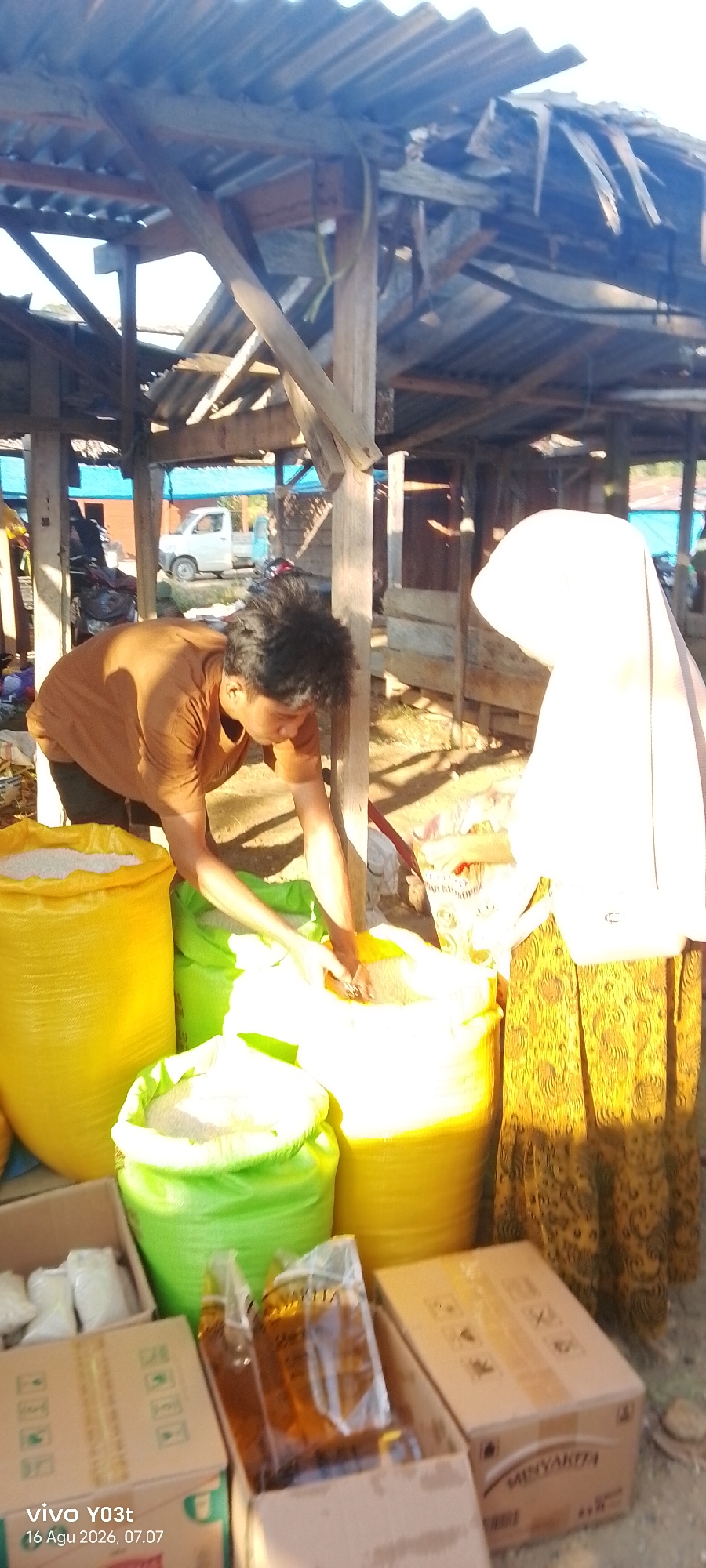 In Palosilae, Indonesia, a market stall under a weathered, partially collapsed wooden structure displays large sacks of rice. A man with dark, curly hair, wearing a brown t-shirt, is reaching into a large yellow sack, appearing to be measuring or scooping rice. To his right, a person wearing a light-colored hijab and a patterned yellow skirt is standing, their back to the camera. In the foreground, cardboard boxes contain smaller bags of what appears to be rice and bottles of cooking oil. A white pickup truck is visible in the background, along with other market structures and foliage. The scene is illuminated by bright sunlight, casting shadows on the ground.