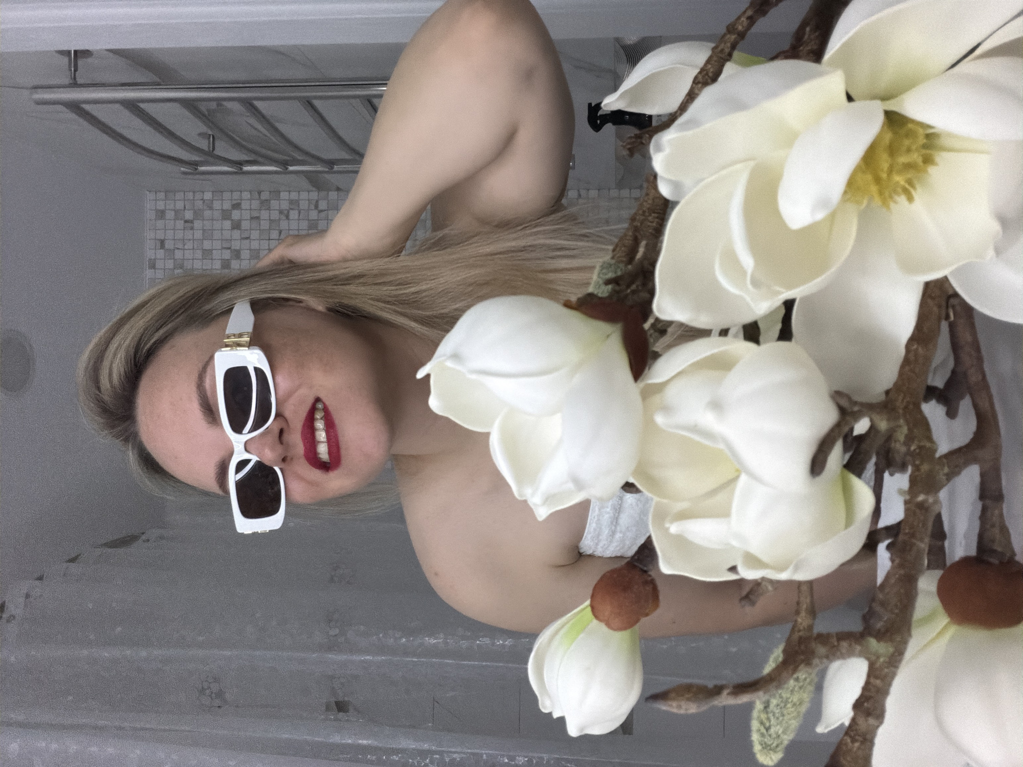 A woman with blonde hair and red lipstick is lying in a bathtub. She is wearing white-framed sunglasses. A branch of artificial white flowers is positioned in the foreground. The bathroom has a tiled wall and a towel rack. The location is not specified. The city is Votkinsk, Russia.