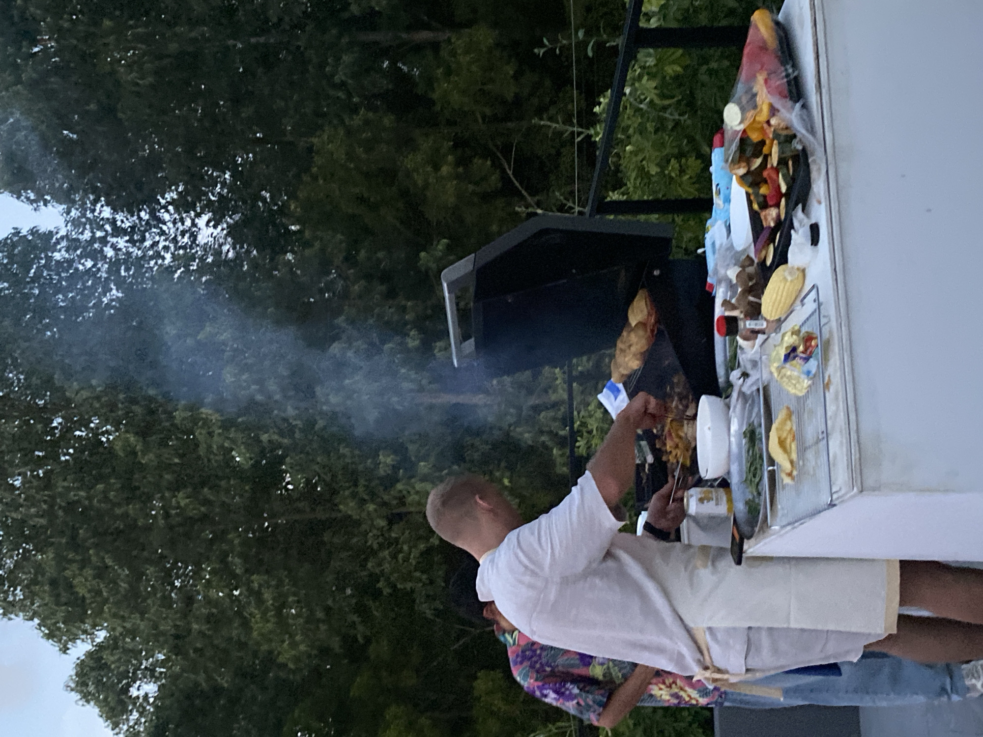 This outdoor image depicts a lively grilling scene, likely during the late afternoon or early evening, given the soft lighting and visible smoke against the foliage. A man, positioned with his back to the camera, is the central figure, actively tending a black barbecue grill. He is dressed in a white t-shirt and light-colored shorts, using tongs to manage items on the grill, specifically skewers that appear to be loaded with meat or vegetables. Plumes of smoke billow upwards from the grill, indicating active cooking.
Another individual, partially obscured behind the main griller, is visible wearing a brightly patterned, colorful shirt. To the right of the grill, a white outdoor counter or table is organized with various food preparations. Notable items include trays of colorful vegetable skewers, possibly featuring bell peppers, zucchini, and onions, along with corn on the cob, bowls, and other containers, suggesting a range of items ready for cooking or serving.
The background is filled with a dense canopy of lush green trees, hinting at a tropical or verdant environment, consistent with the Choeng Thale, Thailand, location context. The overall atmosphere appears casual and focused on meal preparation in an outdoor setting. No discernible text is visible in the scene.
