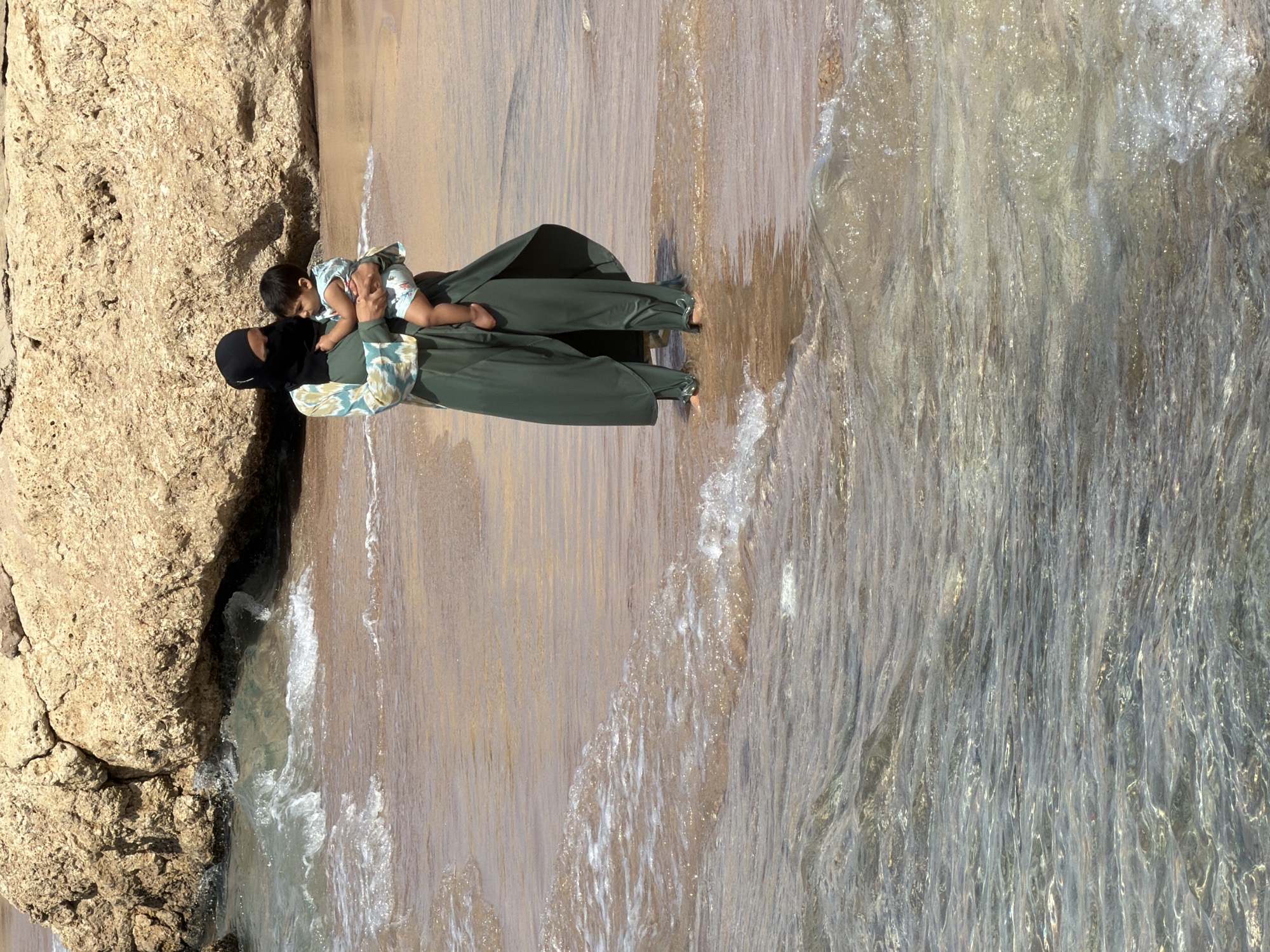 A woman, wearing a black head covering (niqab, covering face except eyes) and a long, dark green dress with patterned sleeves, is depicted holding a baby. The baby, dressed in a light-colored, patterned romper, is nestled against the woman's chest, looking towards her.
They are standing on a wet, sandy area at the edge of shallow, clear water. To their left is a large, rugged rock formation of light brown or beige color. The translucent water, showing hints of the sandy bottom, extends to their right, with small ripples and waves visible around their bare feet. Bright sunlight creates highlights and reflections on the water's surface.
The scene captures a serene moment between the woman and baby by the sea, suggesting affection and comfort. The bright lighting indicates it is daytime, likely a sunny and warm day. The natural elements of rock, sand, and clear water place the setting at a coastal beach or shore. The location context provided specifies Sharm El Sheikh, Egypt. No visible text is present in the image.