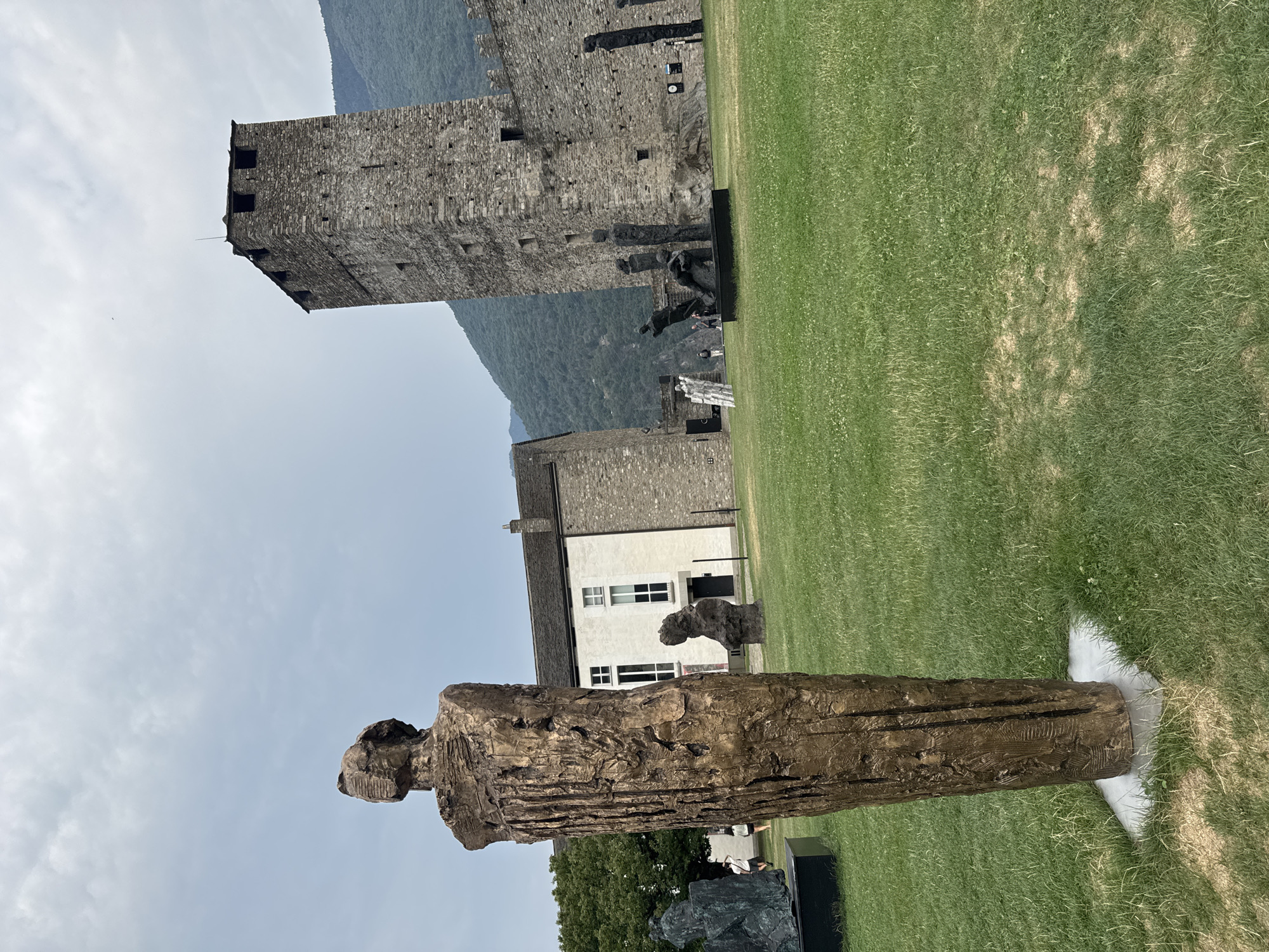 An outdoor sculpture garden is situated in front of a medieval stone castle. In the foreground, a tall, textured bronze sculpture of a human figure stands on a white base. To the left, a dark green sculpture rests on a black rectangular plinth. In the midground, several abstract dark metal sculptures of human figures are arranged on the grass. Behind these, a white building with a dark roof is visible, adjacent to the castle walls. The castle features a tall square tower and a long, fortified wall constructed from rough-hewn stones. The sky is overcast and grey. The location is Castel Grande in Bellinzona, Switzerland.