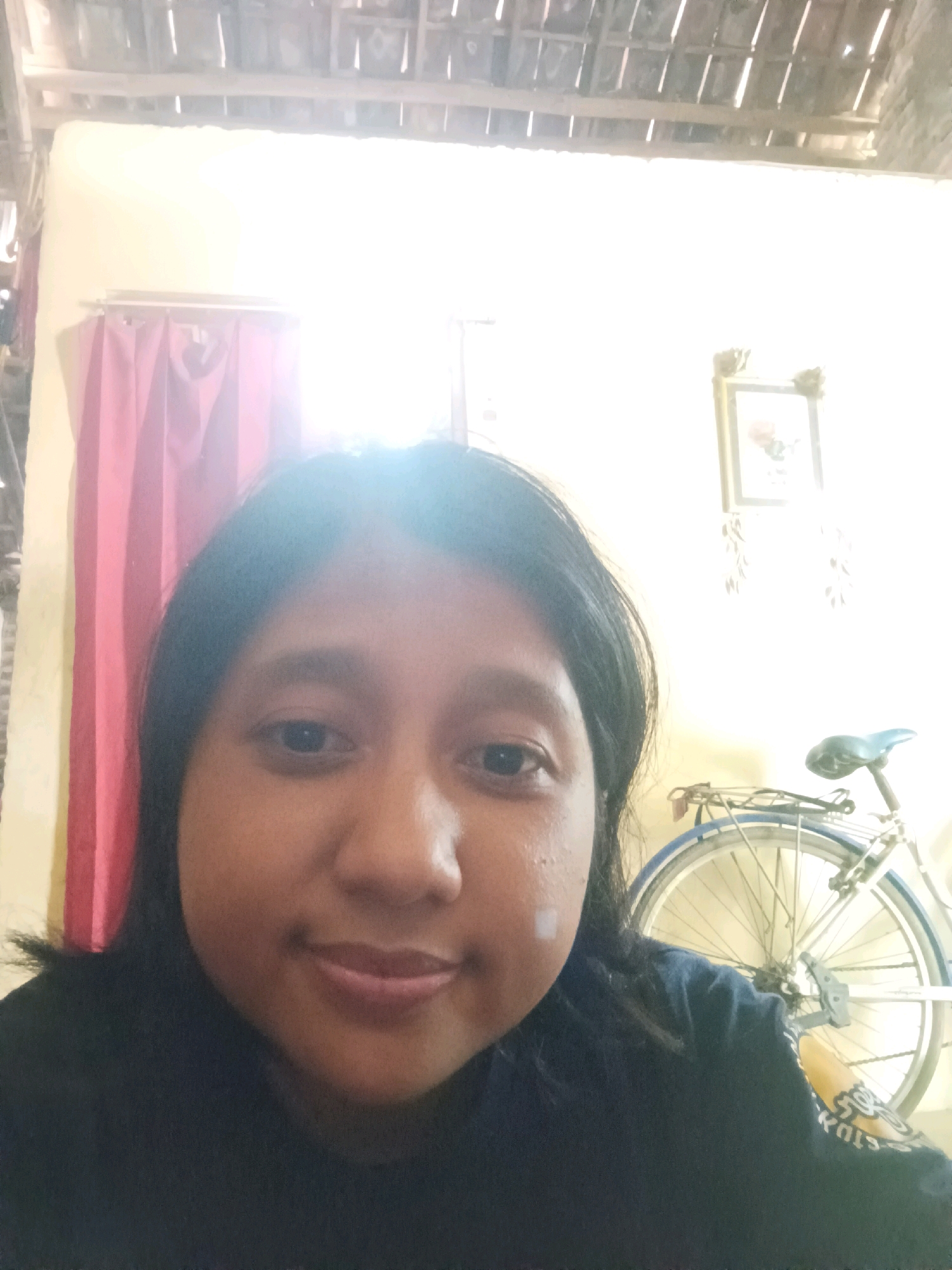 A selfie is taken from a low angle, capturing a woman with dark hair and a neutral expression. She is wearing a dark blue shirt. Behind her, a pink curtain hangs to her left. To her right, a bicycle with a blue seat and a white rim is visible, along with a framed picture on the wall. The ceiling, made of exposed wooden beams, is visible at the top of the image. The setting appears to be indoors, possibly a home in Canga'an, Indonesia, given the style of the ceiling and the presence of traditional elements like the curtain. The lighting is bright and washed out, suggesting the image was taken during the day with strong natural light.