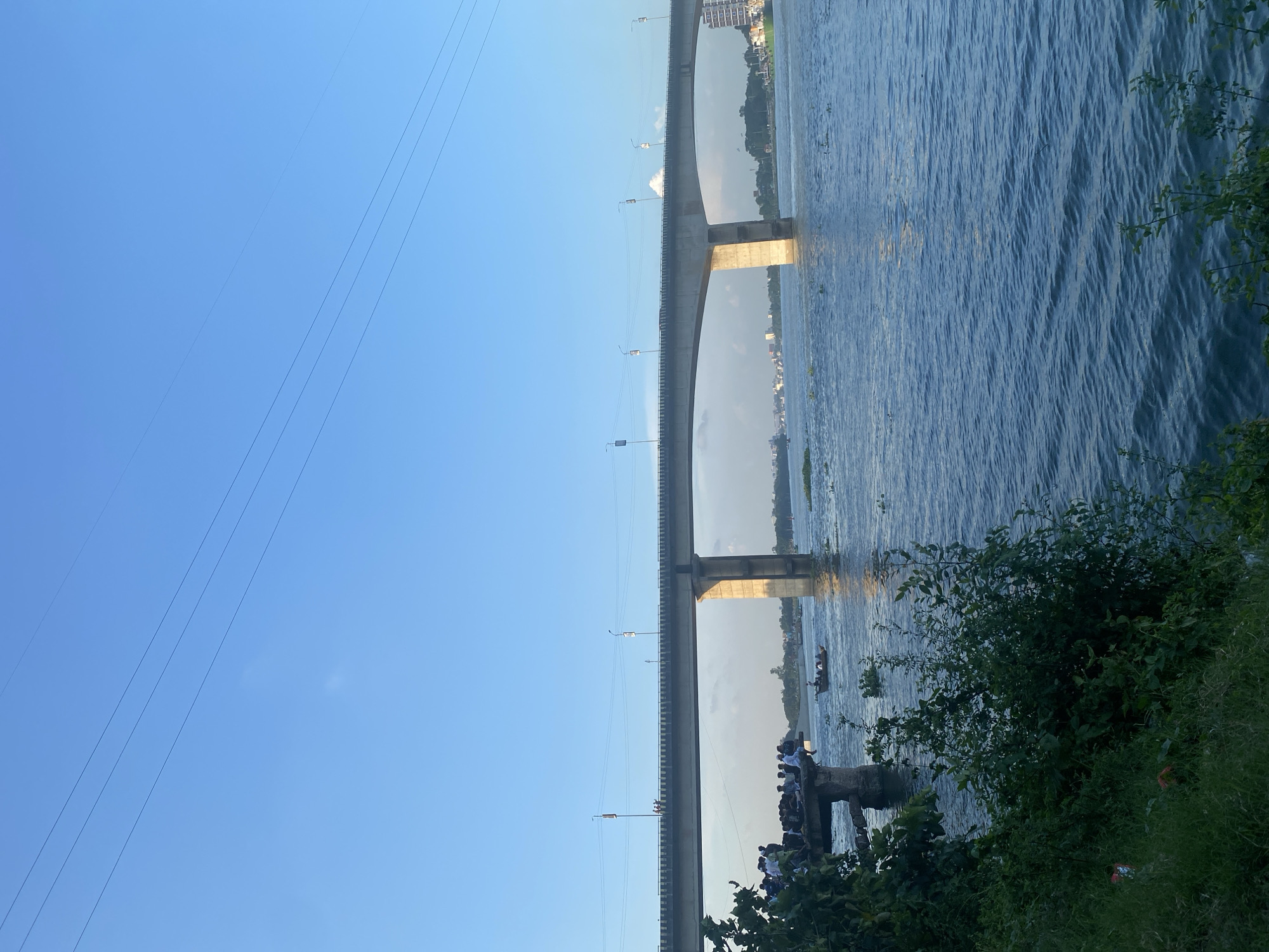 The image depicts a view of the Buriganga River in Dhaka, Bangladesh, with a long concrete bridge spanning across it. The bridge has multiple pillars supporting its structure and is illuminated by streetlights. On the left side of the image, a group of people are gathered on a concrete structure overlooking the river. A small boat with people on board is seen in the water near this gathering. The river itself is calm, with some floating vegetation visible. The sky is clear and blue, with a few power lines visible across the top of the frame. In the background, the city skyline is visible, with buildings and trees lining the opposite bank of the river. The overall scene suggests a public area where people are observing the river and the bridge, possibly during the late afternoon or early evening.