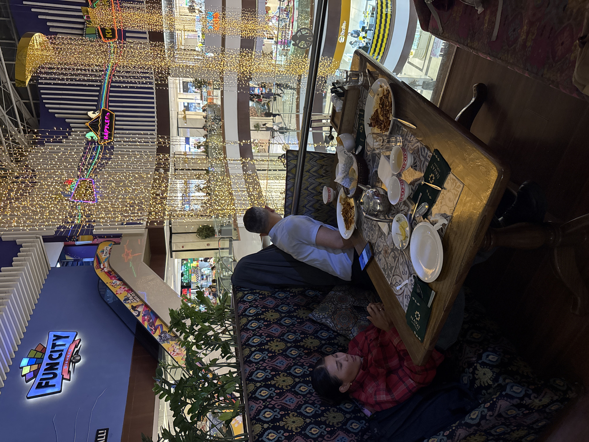 The image captures an indoor scene within a multi-story shopping mall or entertainment complex in Bishkek City, Kyrgyzstan, likely during the evening due to the pervasive artificial lighting. The foreground features a restaurant table where two individuals are present. A man, seen from behind, sits at the table, wearing a light-colored shirt. A woman is reclining on a patterned sofa, wearing a red plaid shirt, appearing relaxed.
On the wooden table, there are plates of food, a teapot, traditional-style tea cups, and drinking glasses, suggesting a meal or tea service. A mobile phone also rests on the table. A menu or booklet with the text "NAVAT KARLAHA" is visible, identifying the establishment as likely a branch of the "Navat" restaurant chain.
The background showcases the bustling mall environment, adorned with numerous strings of warm, golden lights that create a festive atmosphere. Various mall levels, escalators, and storefronts are visible. Prominent signage includes "FUNCITY BISHKEK" on a blue wall, indicating an entertainment area. Another sign reads "КАРУСЕЛЬ" (KARUSEL), suggesting a carousel ride. A "Beeline" logo is also seen, alongside a safety sign in Russian stating "ПОЖАЛУЙСТА ДЕРЖИТЕ ДЕТЕЙ ДАЛЬШЕ ОТ ПЕРИЛ" (PLEASE KEEP CHILDREN AWAY FROM RAILINGS). The overall scene depicts a blend of dining and leisure activities within a vibrant mall setting.