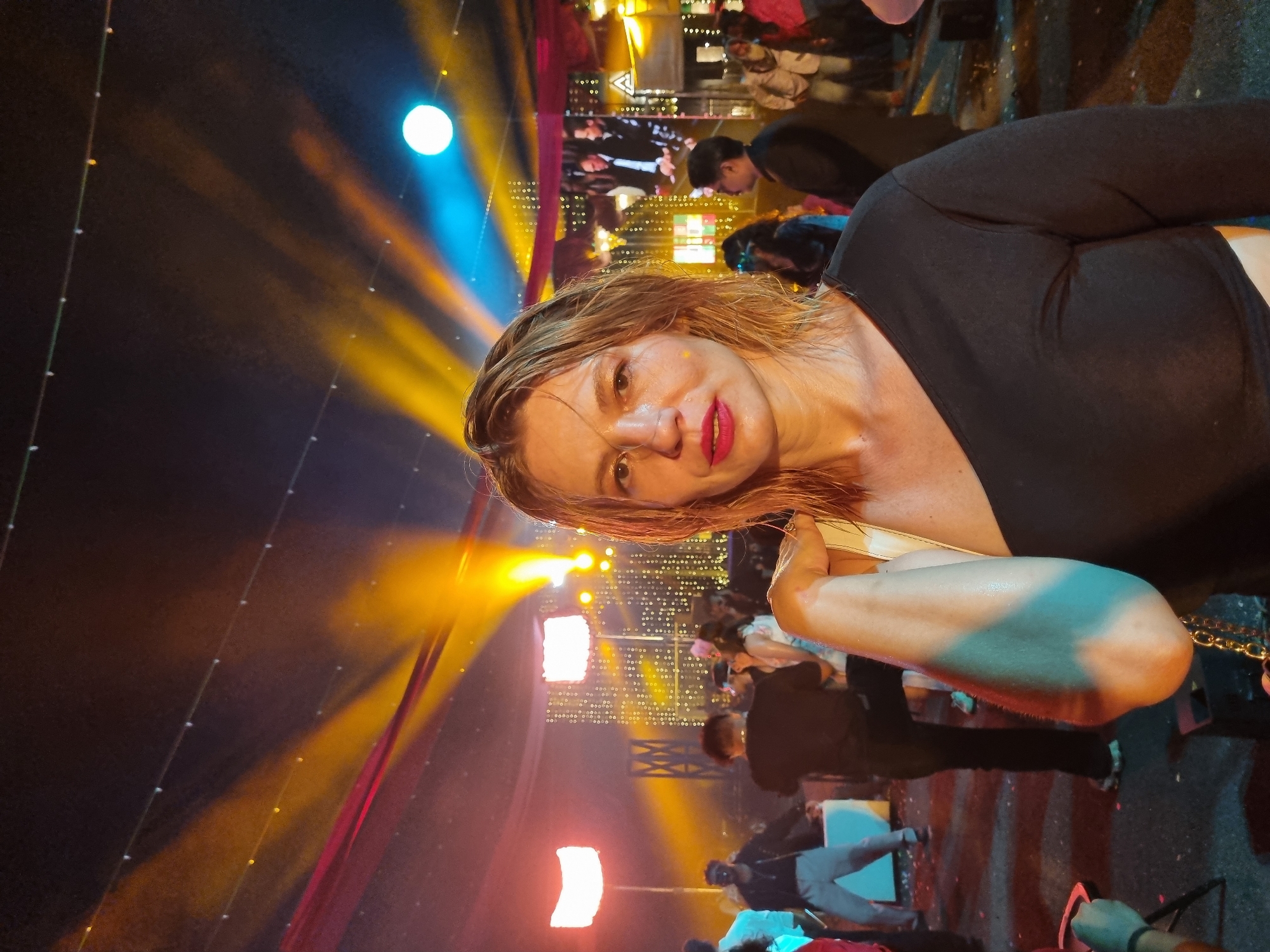 A portrait-oriented image, captured at night, features a woman in the foreground within a vibrant, artificially lit event setting. The woman, with light hair and fair skin, directly faces the camera, sporting red lipstick and a black top. Her face and hair appear damp, possibly from sweat or humidity, with a noticeable sheen. She holds a light-colored bag strap with her right hand, part of which is visible in the lower right.
The background is a dynamic scene illuminated by numerous colorful lights. Beams of yellow and blue light emanate from sources above, casting a festive glow across the dark space. String lights with small white bulbs are visible across the upper portion of the frame. In the mid-ground, various people are present, mostly indistinct and blurred, suggesting a crowd. Some bright white rectangular light panels are also visible. The overall atmosphere suggests a lively party, concert, or outdoor festival. The setting, as per provided context, is Kalamassery, India. There is no visible text in the image.
