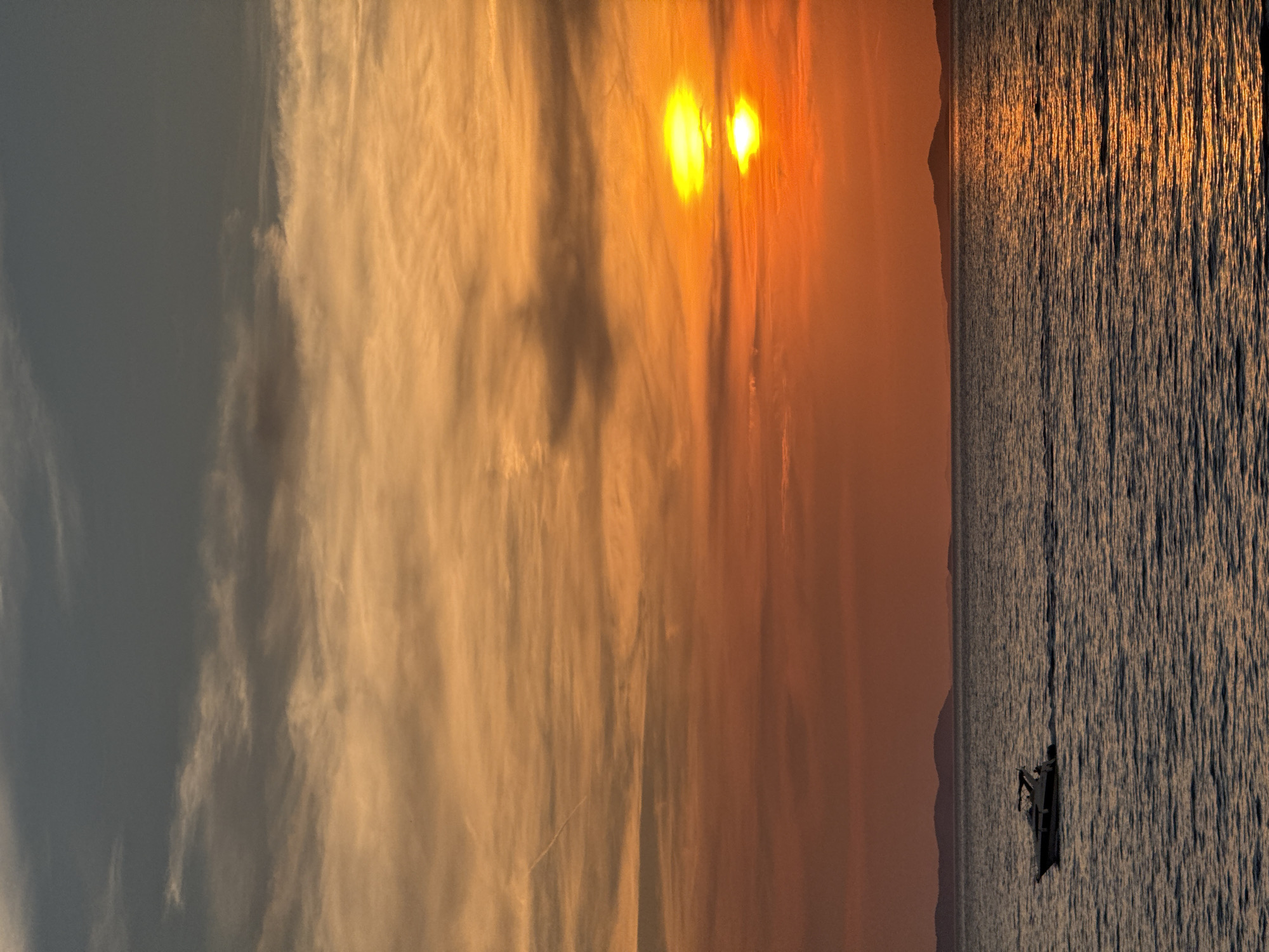 This image captures a sunset over the Adriatic Sea, with the silhouette of the Croatian coast and islands in the distance. The sky transitions from a deep blue at the top to vibrant oranges and yellows near the horizon, painted with wispy clouds. The sea below is a deep blue, reflecting the warm hues of the sunset. A small motorboat, dark against the water, is moving from left to right, leaving a faint wake. The scene is set in Primošten, Croatia, known for its picturesque coastal landscapes.
