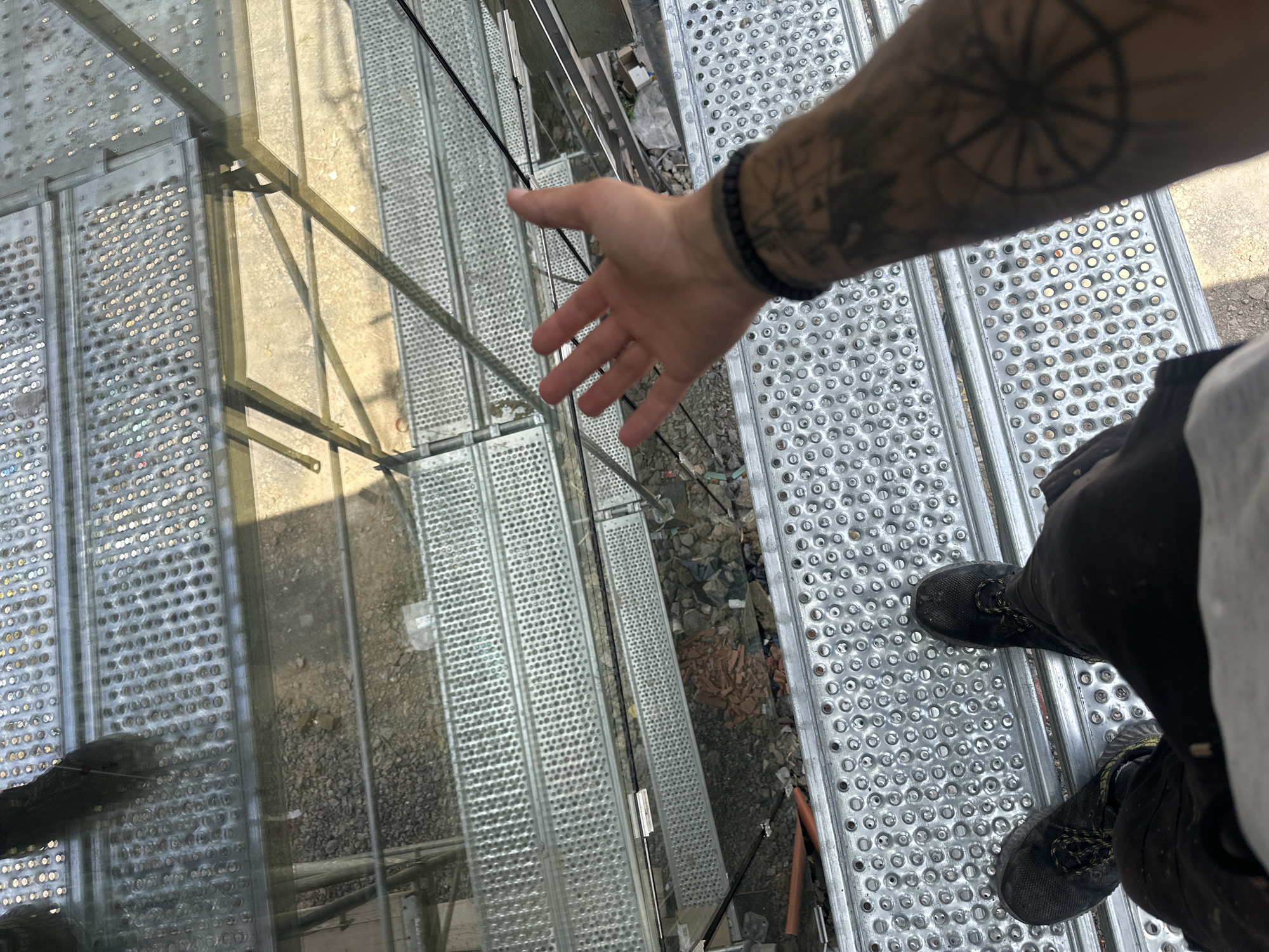 The image shows a first-person perspective of a person standing on a metal grate walkway, likely part of a construction or industrial site. The walkway is made of perforated metal planks, with a reflective surface that shows shadows and light patterns. Below the walkway, visible through the grates and a transparent surface, is a ground covered with gravel and debris.
In the foreground, the person's hand is extended, palm open, as if reaching out or testing the space. The person is wearing dark pants and black athletic shoes with yellow laces. The person's left arm, with tattoos including what appears to be a compass rose, is also visible, adorned with a beaded bracelet.
The scene suggests an outdoor environment, with daylight illuminating the metal structures. The reflections indicate a sunny day. The presence of construction materials and the overall industrial aesthetic point to a location under development or maintenance. The perspective from above the ground implies the walkway is elevated. The location is identified as Stara Zagora, Bulgaria. No visible text is present in the image.