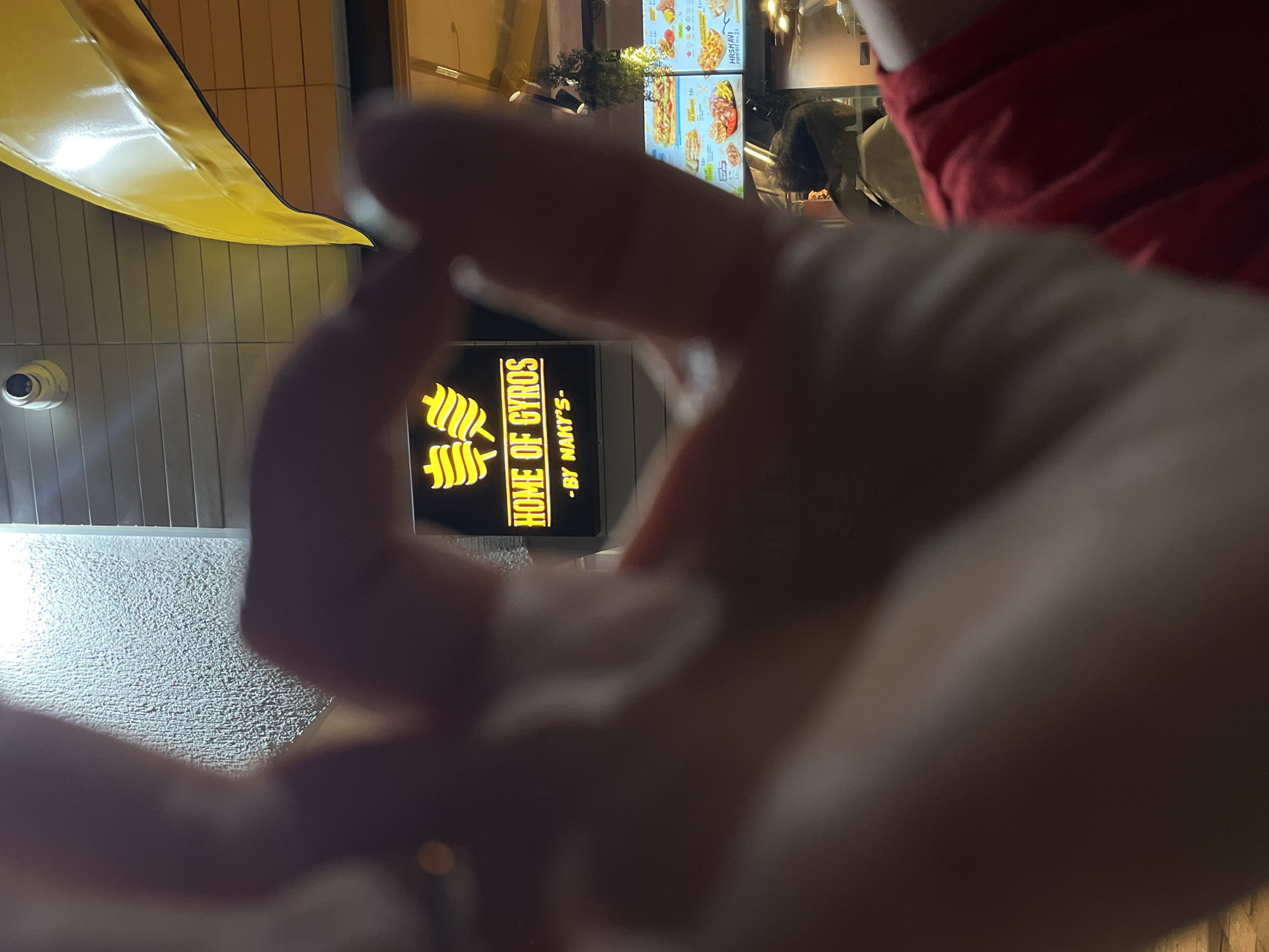 The image shows a close-up of a person's hand, with their fingers forming an "ok" gesture in the foreground, obscuring a neon sign that reads "HOME OF GYROS - BY NAKY'S". The sign is yellow and black, with stylized images of gyros. Behind the sign, a menu board displays various food items and prices. A yellow awning is visible at the top of the frame. The scene appears to be outdoors at night, possibly in a street food establishment in Budva, Montenegro, as suggested by the details in the broader context of the image. The lighting is dim, with the neon sign and menu board being the primary light sources. The background is out of focus, but suggests the presence of other people and structures.