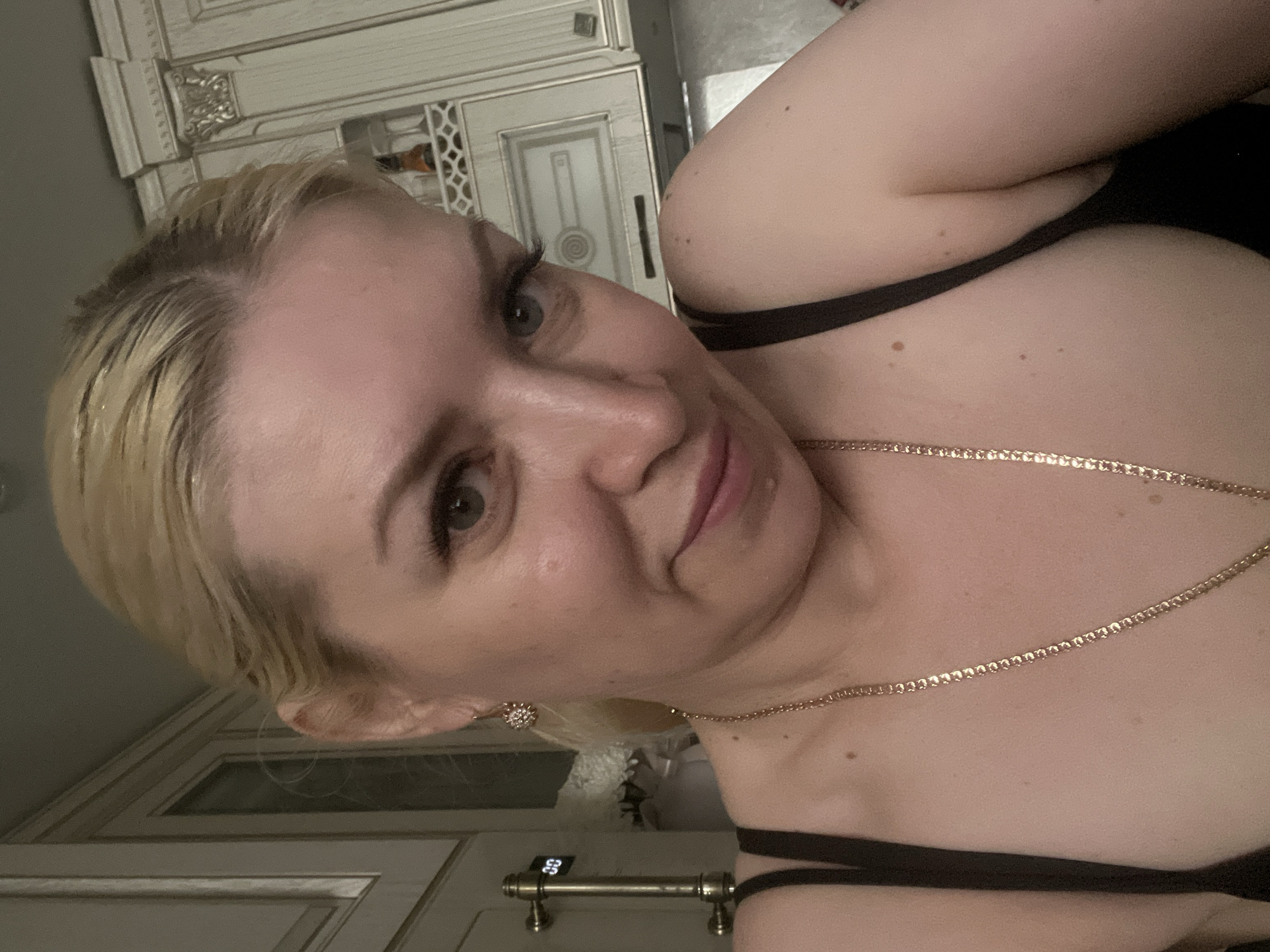 The image shows a close-up, selfie-style portrait of a blonde woman with blue eyes. She is wearing a black camisole and a gold necklace. Her hair is pulled back. She is looking towards the camera with a slight smile. The background appears to be a kitchen with light-colored cabinets. There is a digital clock visible on the left side of the image displaying "00". There are no indications of the specific location within the city of Tyumen, Russia.