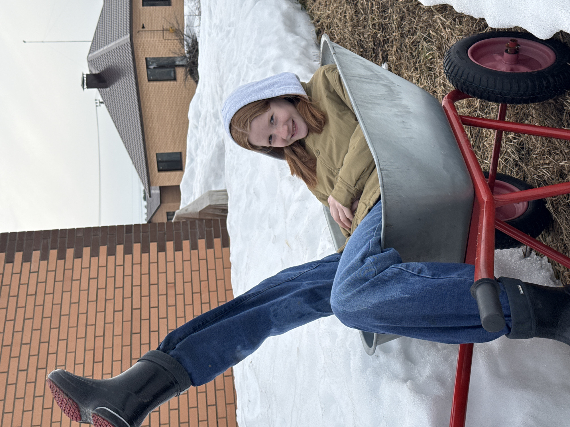 An outdoor scene in Chaikovsky, Russia, depicts a young female seated within a silver-colored wheelbarrow featuring a red frame and red-centered wheels. The female has reddish-brown hair, wears a light grey hoodie over a tan jacket, blue jeans, and black rubber boots. Her left leg is extended vertically upwards, displaying the red sole of the boot and the "Nordman" brand on the shaft. She is smiling and looking directly at the viewer. The ground consists of melting snow patches interspersed with dry, brown grass and dirt. In the background, a two-toned brick building is visible on the left, and another single-story brick building with dark windows, a dark brown tiled roof, and a black chimney is present on the right, under an overcast sky. Overhead power lines are visible in the distance.