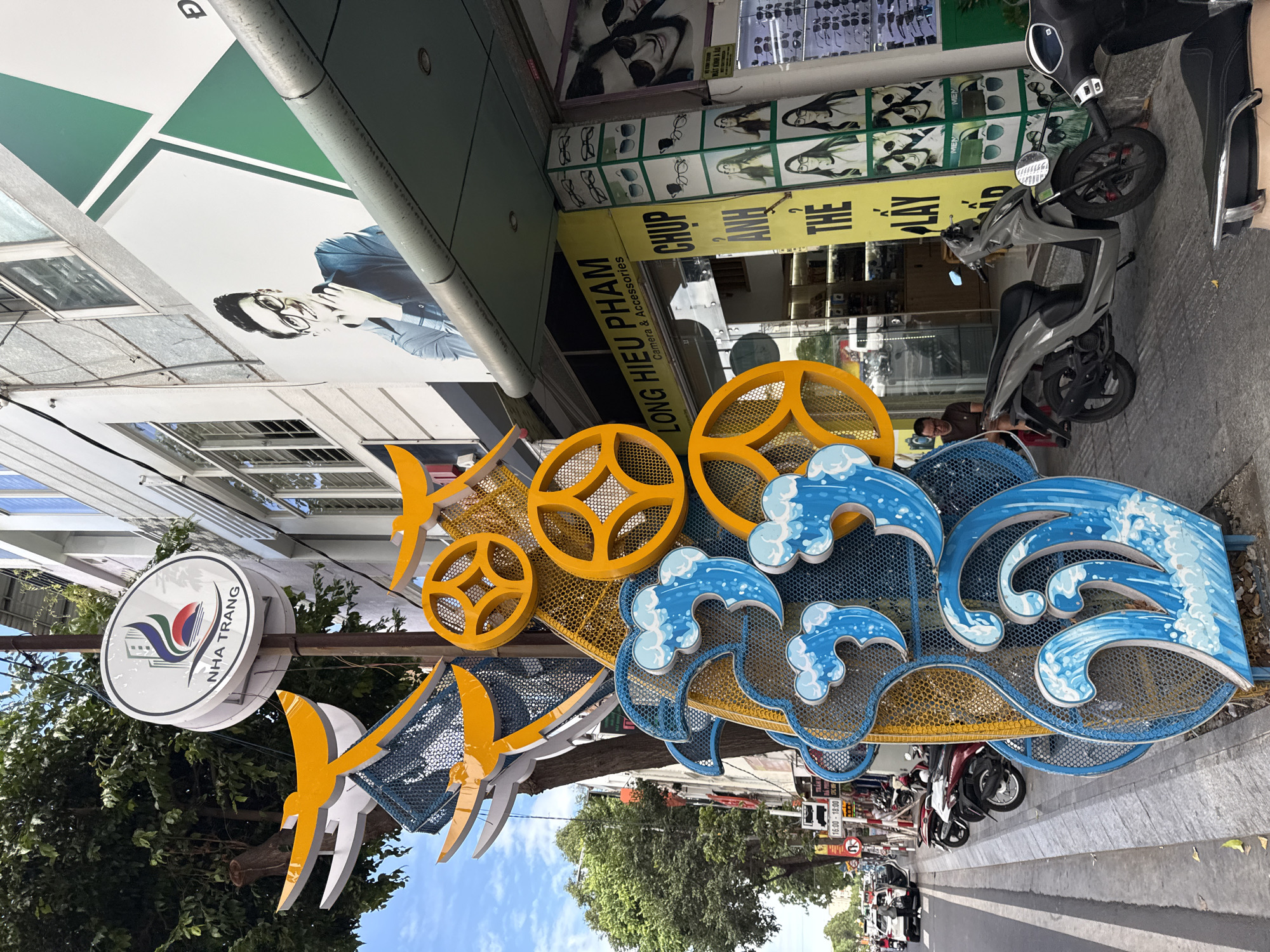 The image depicts a street scene in Nha Trang, Vietnam, featuring a decorative sculpture on the sidewalk. The sculpture is composed of bright yellow and blue elements, resembling stylized waves and wheels. To the right of the sculpture is the facade of a shop with a yellow sign that reads "LONG HIEU PHAM Camera & Accessories." Above the shop entrance, a large banner displays a young man wearing glasses. Further to the right, another shop window displays various eyewear products. In the background, a tree with green foliage is visible, and a few motorcycles are parked along the street. The sky above is partly cloudy and blue. A few people are present, including a man sitting on a red stool near the shop entrance.