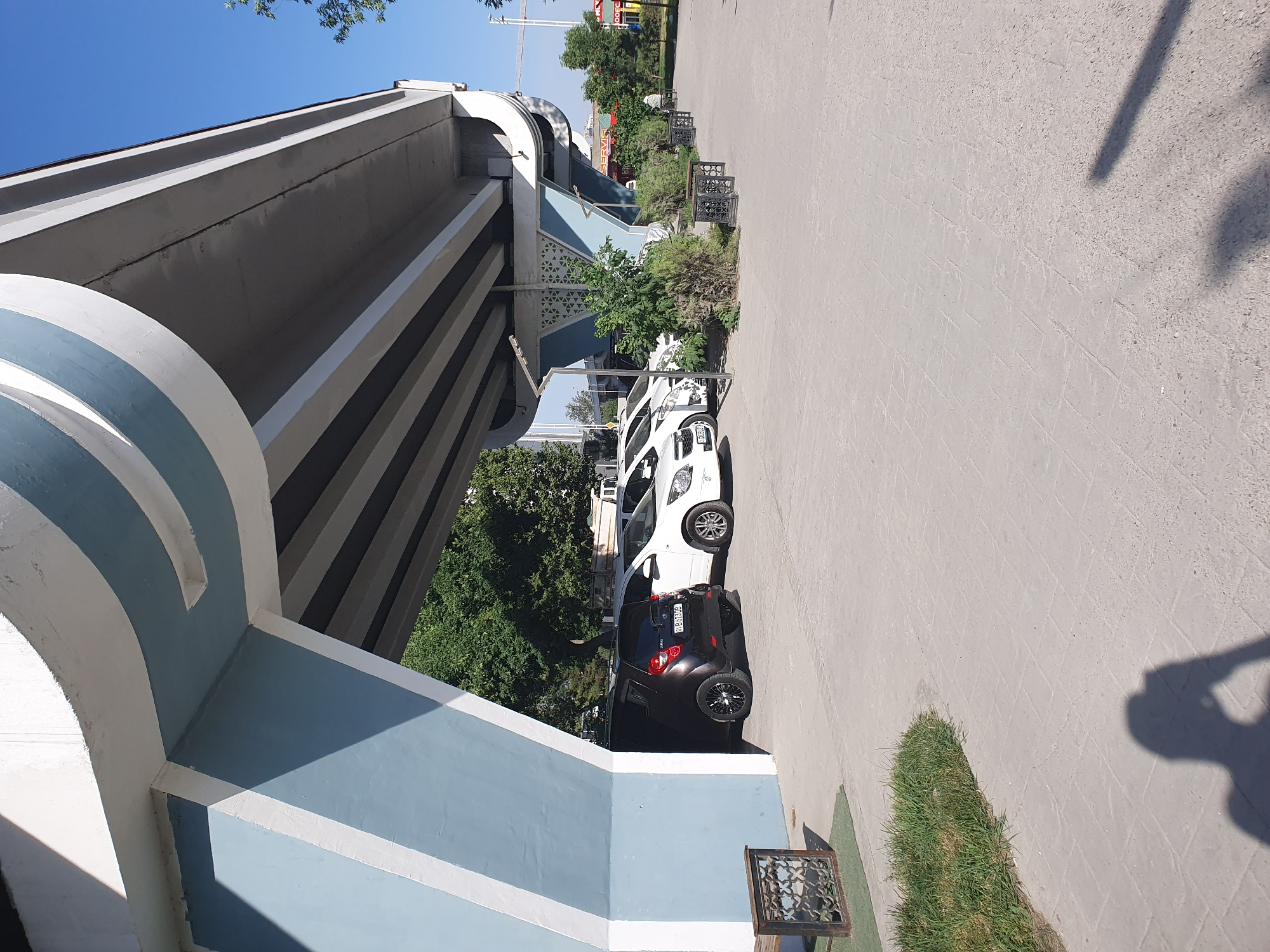 Underneath a blue and white painted overpass in Tashkent, Uzbekistan, a row of parked cars is visible. The cars include a dark colored compact car in the foreground and several white cars behind it. The ground is paved with square tiles. Lush green trees and bushes are present on the right side of the image, along with benches and decorative planters. The sky is clear and blue.