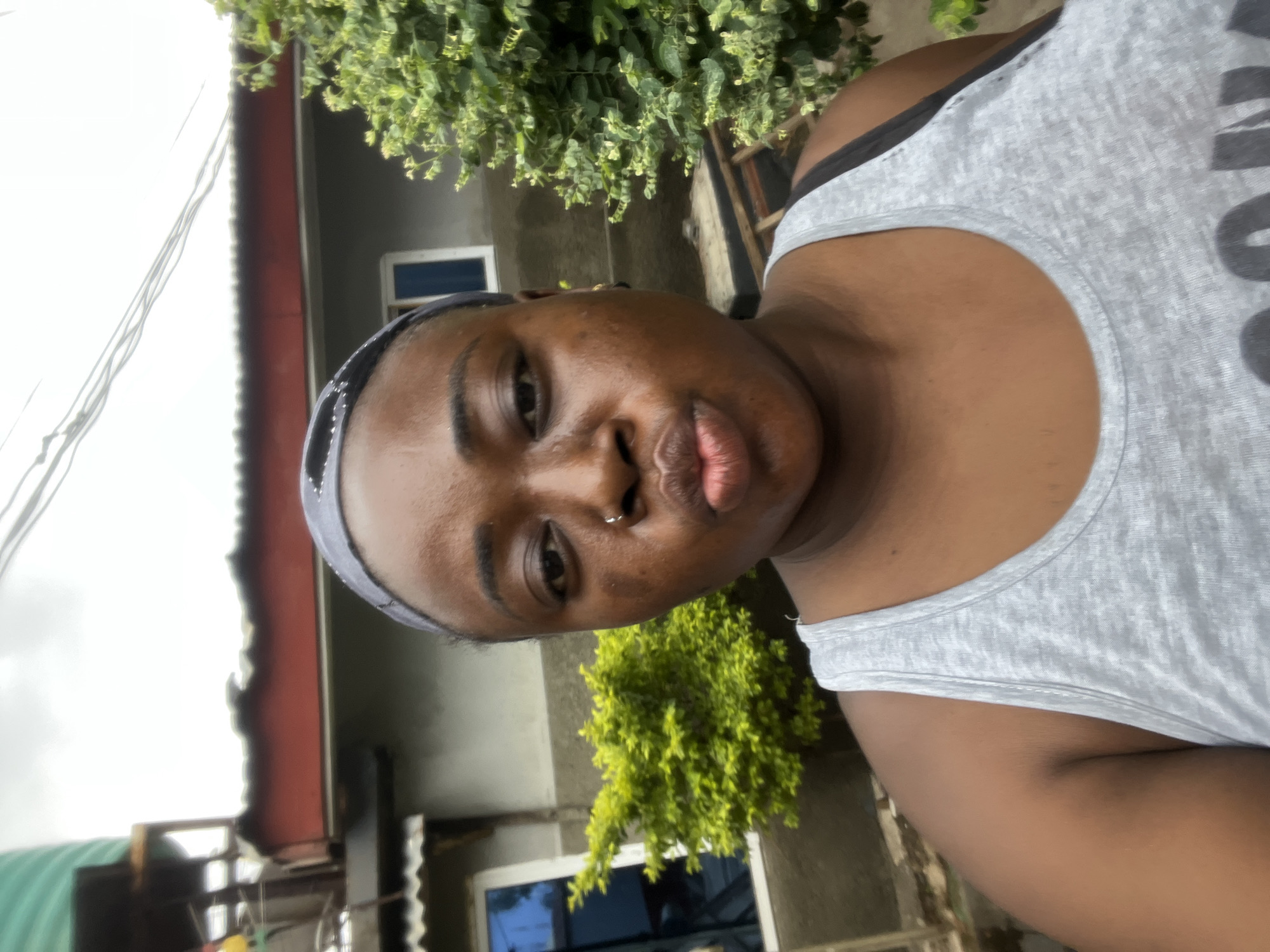 A medium shot captures a person with brown skin wearing a gray tank top and headband. The person has a nose ring and is looking directly at the camera. Behind them, there is a building with gray siding and windows, partially obscured by greenery. There are also power lines visible in the background. The location appears to be outdoors, potentially in a residential area of Jos, Nigeria, although this location is inferred based on the image and provided context, not explicitly identifiable within the image itself.