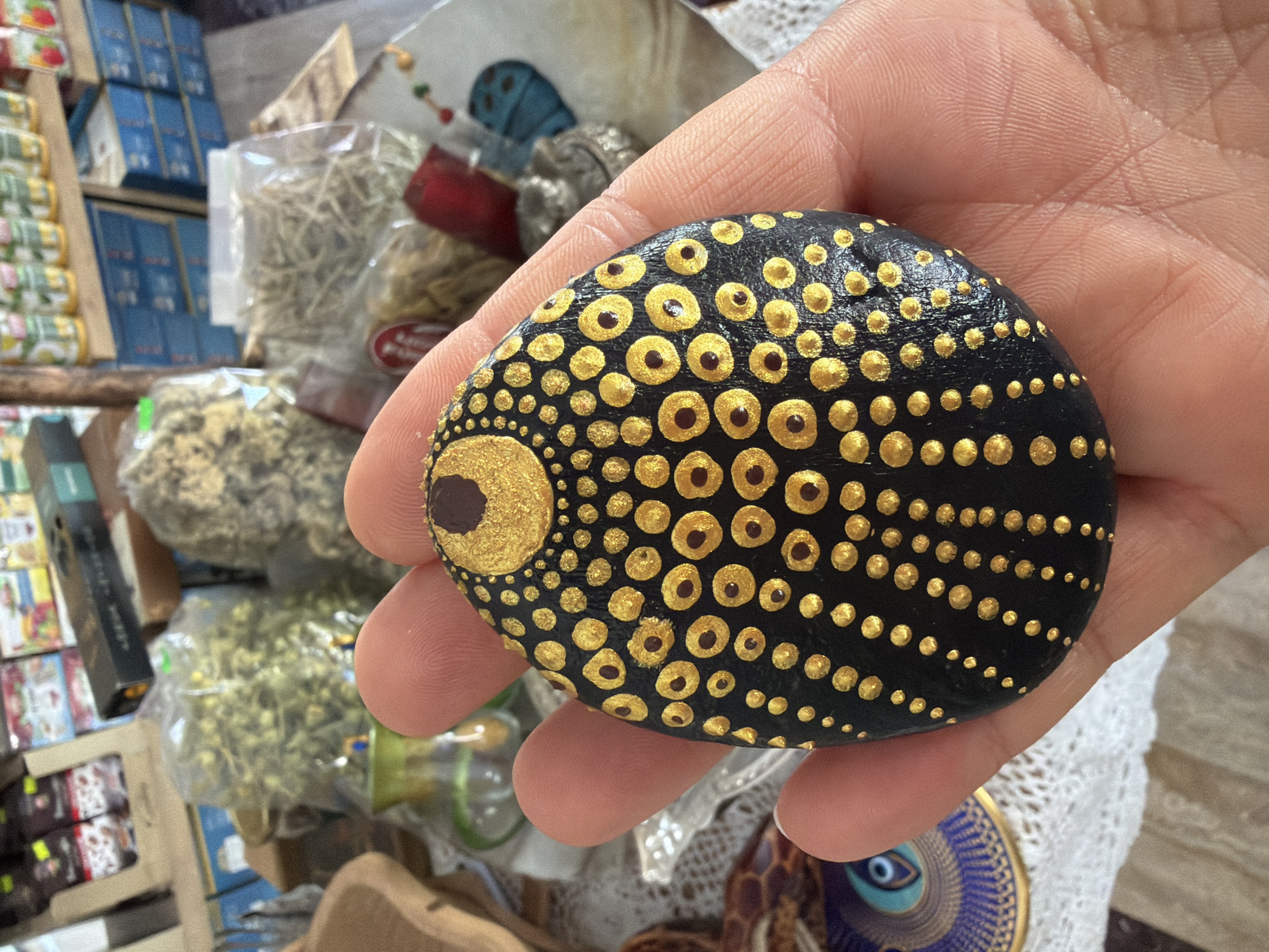 A person's hand holds a black, oval-shaped stone decorated with a mandala pattern of gold and brown dots. The stone is held in the foreground against a background of a market stall. The stall displays various items, including bags of dried herbs, boxes of packaged goods, and other decorative objects. The overall scene suggests a retail environment, possibly a souvenir shop or a market in a tourist area. The specific location is the city of Marmaris, Turkey.