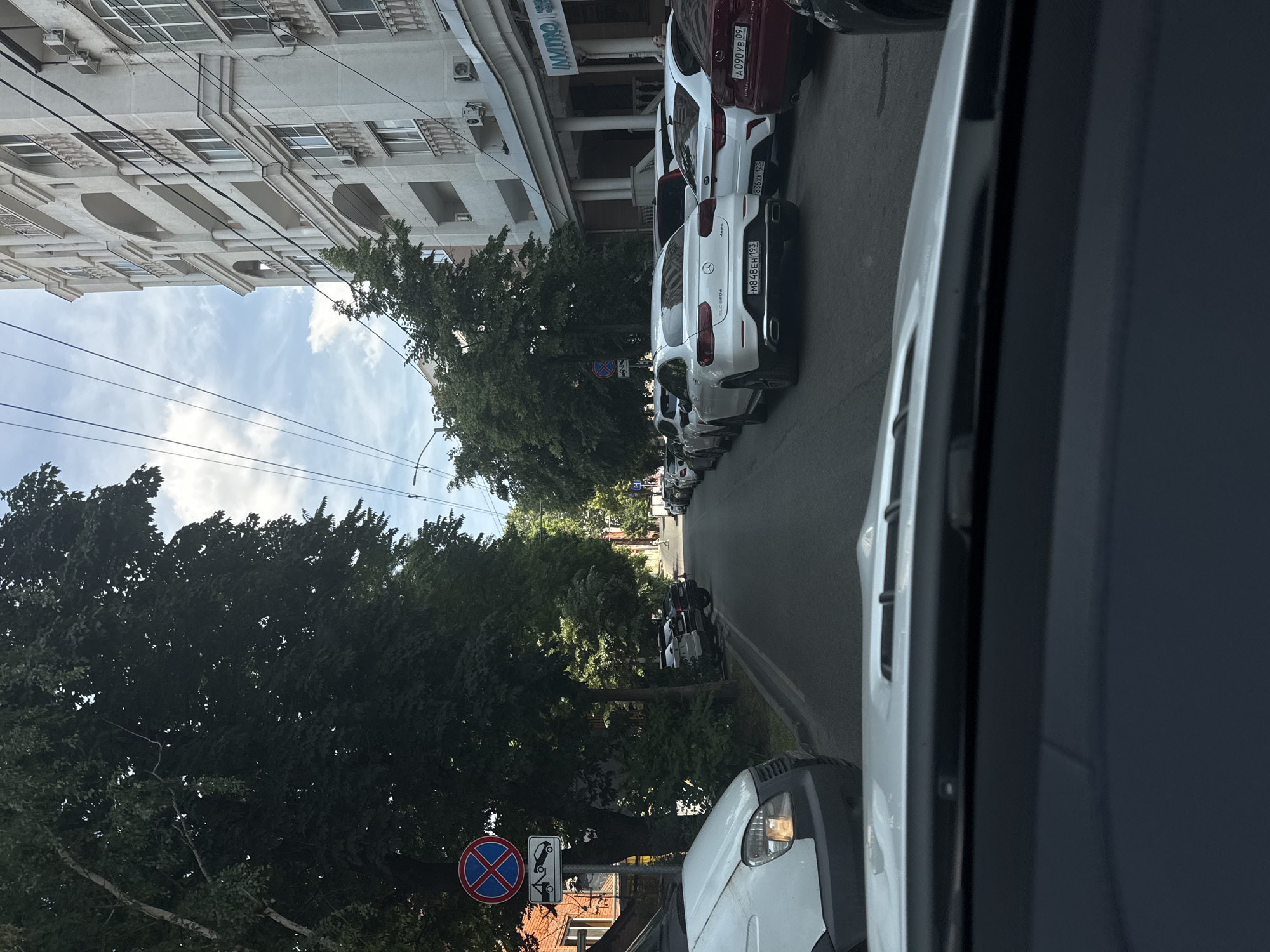 The image depicts a street scene in Krasnodar, Russia, viewed from inside a vehicle. The time of day appears to be daytime, with a bright sky and visible clouds, suggesting fair weather. The street is lined with parked cars on the right side, including several white Mercedes-Benz vehicles. A red car is parked further down the street. On the left side of the street, there is a multi-story building with a light-colored facade, arched windows, and some air conditioning units visible. Trees with lush green foliage are abundant, casting shadows on the street and sidewalk.
Further down the street, more trees and buildings are visible. A street sign with a red circle and a diagonal red bar over a blue background, indicating a prohibition, is present. Another sign below it shows a tow truck icon. The overall impression is of a typical urban environment in a residential or commercial area. There are no people clearly visible in the image. The perspective suggests the vehicle from which the photo is taken is moving or stopped in traffic.
Visible text includes "INVITRO" on a sign on the building. The license plates on the cars are also visible, for example, "MB48EH193" on a white Mercedes. The overall mood is neutral, reflecting an everyday street view.